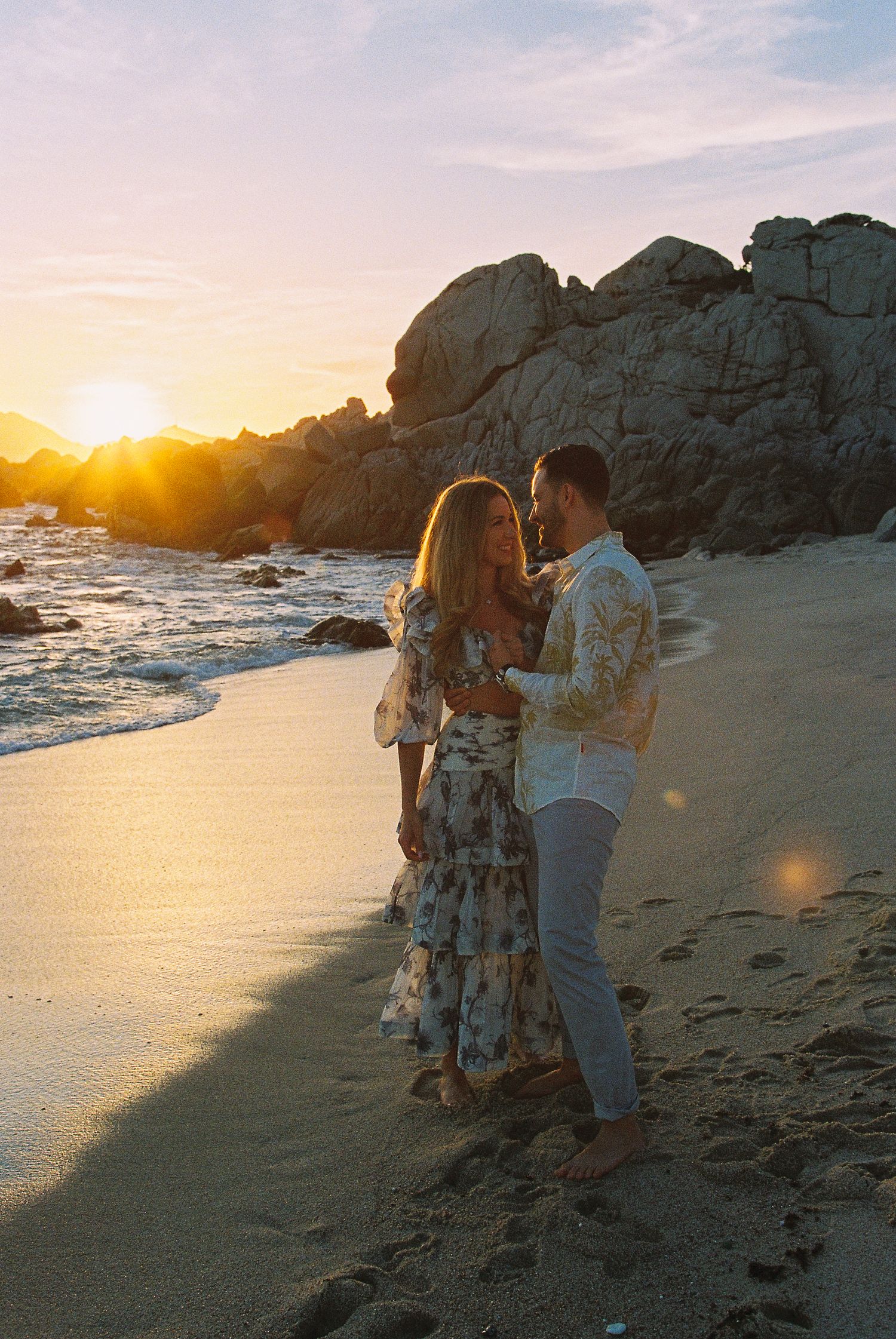 Couple embracing on a sandy beach at golden sunset with rocky cliffs and ocean waves in the background in Cabo San Lucas, Mexico.