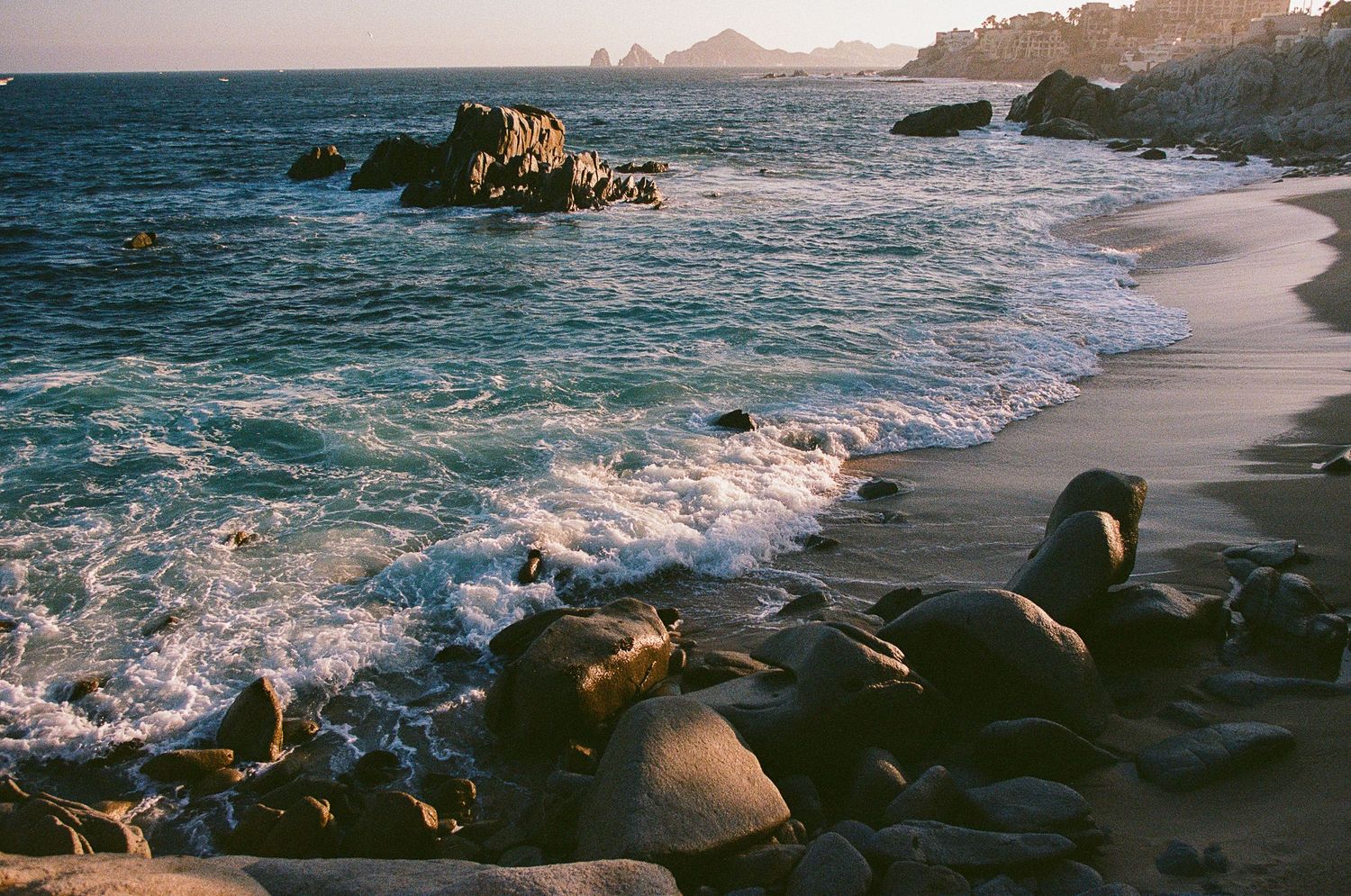 Rocky shoreline with turquoise ocean waves washing onto a sandy beach at golden hour near Cabo San Lucas, Mexico.