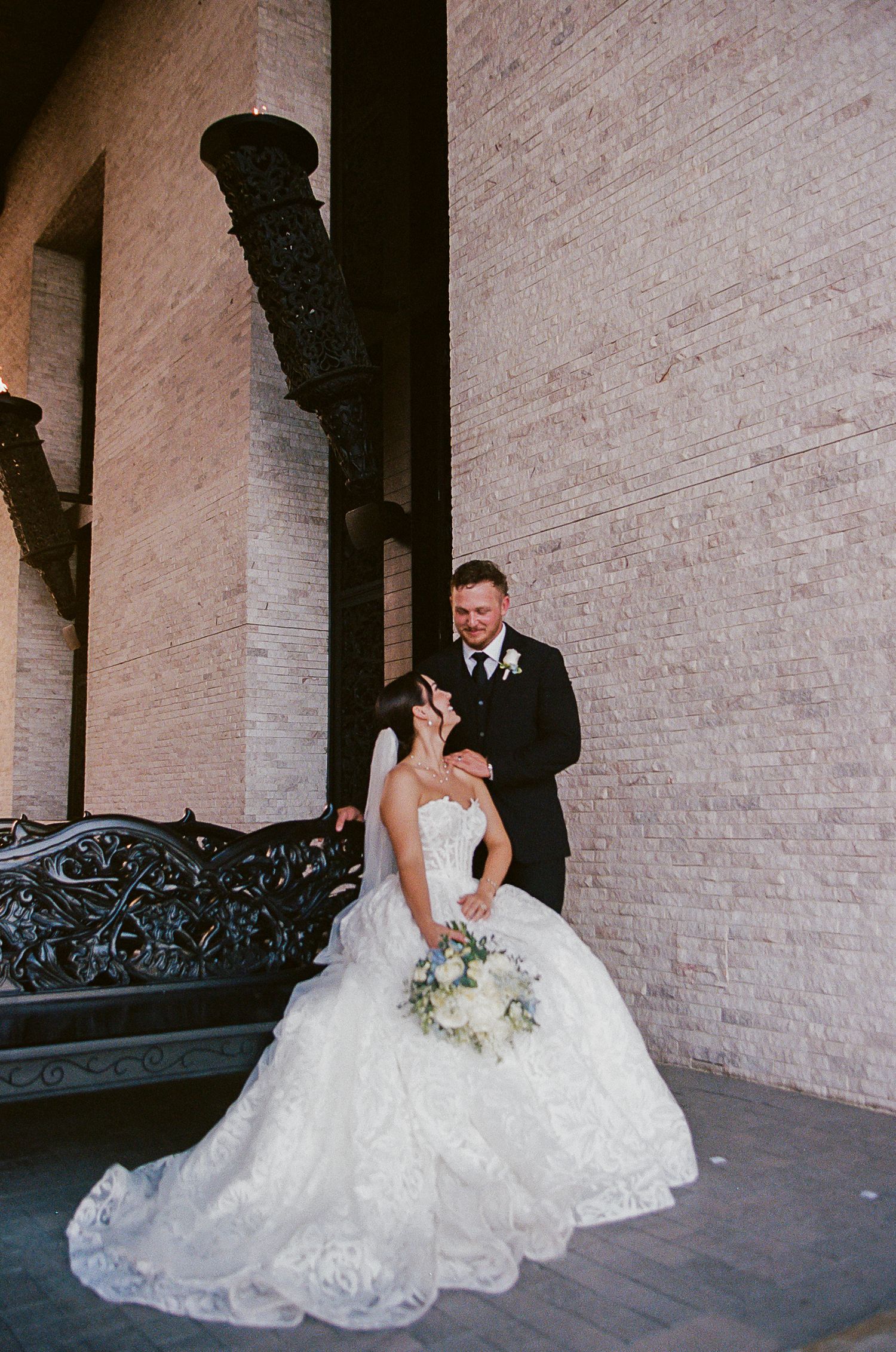 Bride in lace ball gown holding white bouquet poses with groom in black tuxedo against brick wall at elegant wedding venue.