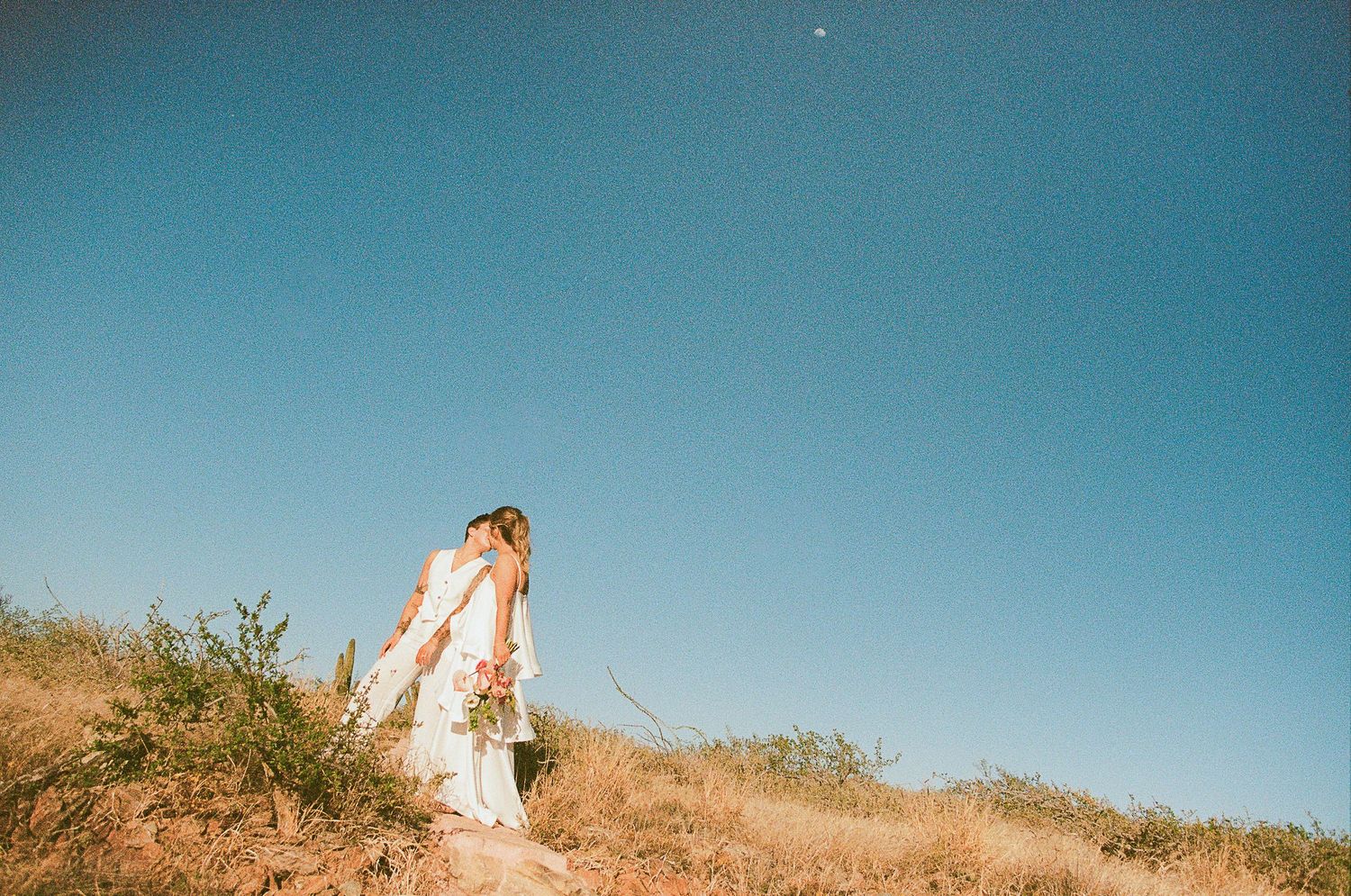 LGBTQ couple kissing under bright blue Baja sky with desert landscape at Hotel San Cristóbal.