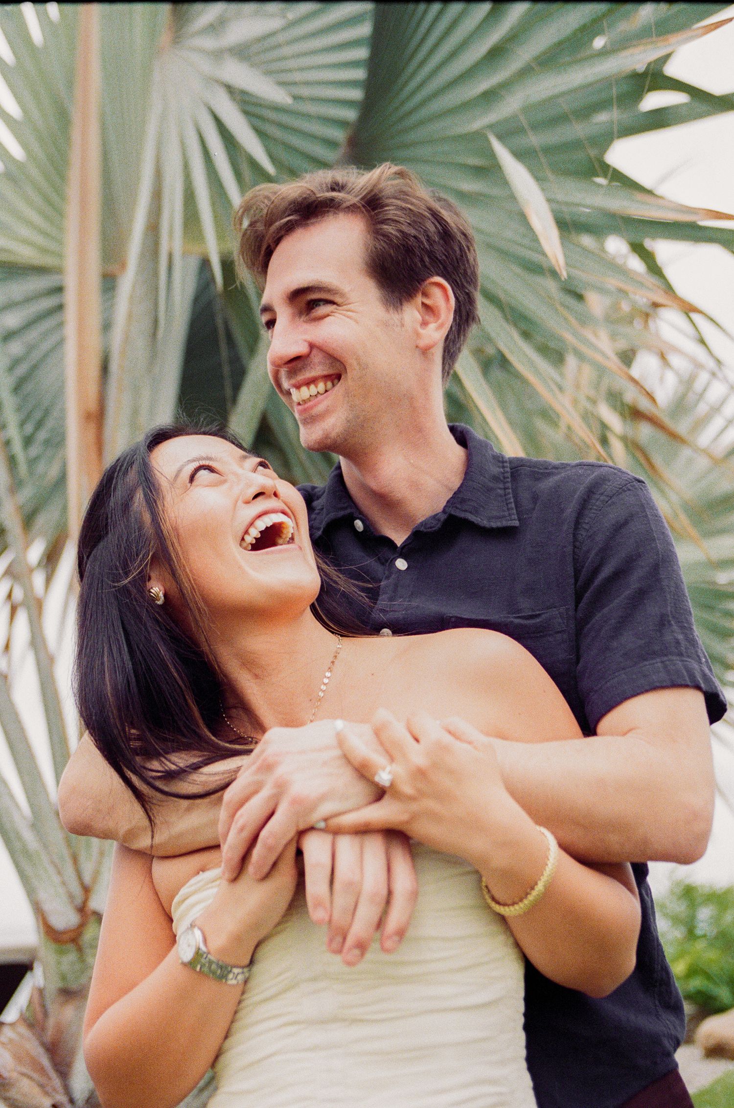 Happy couple embracing and laughing together outdoors near tropical palm leaves in a romantic engagement photo.