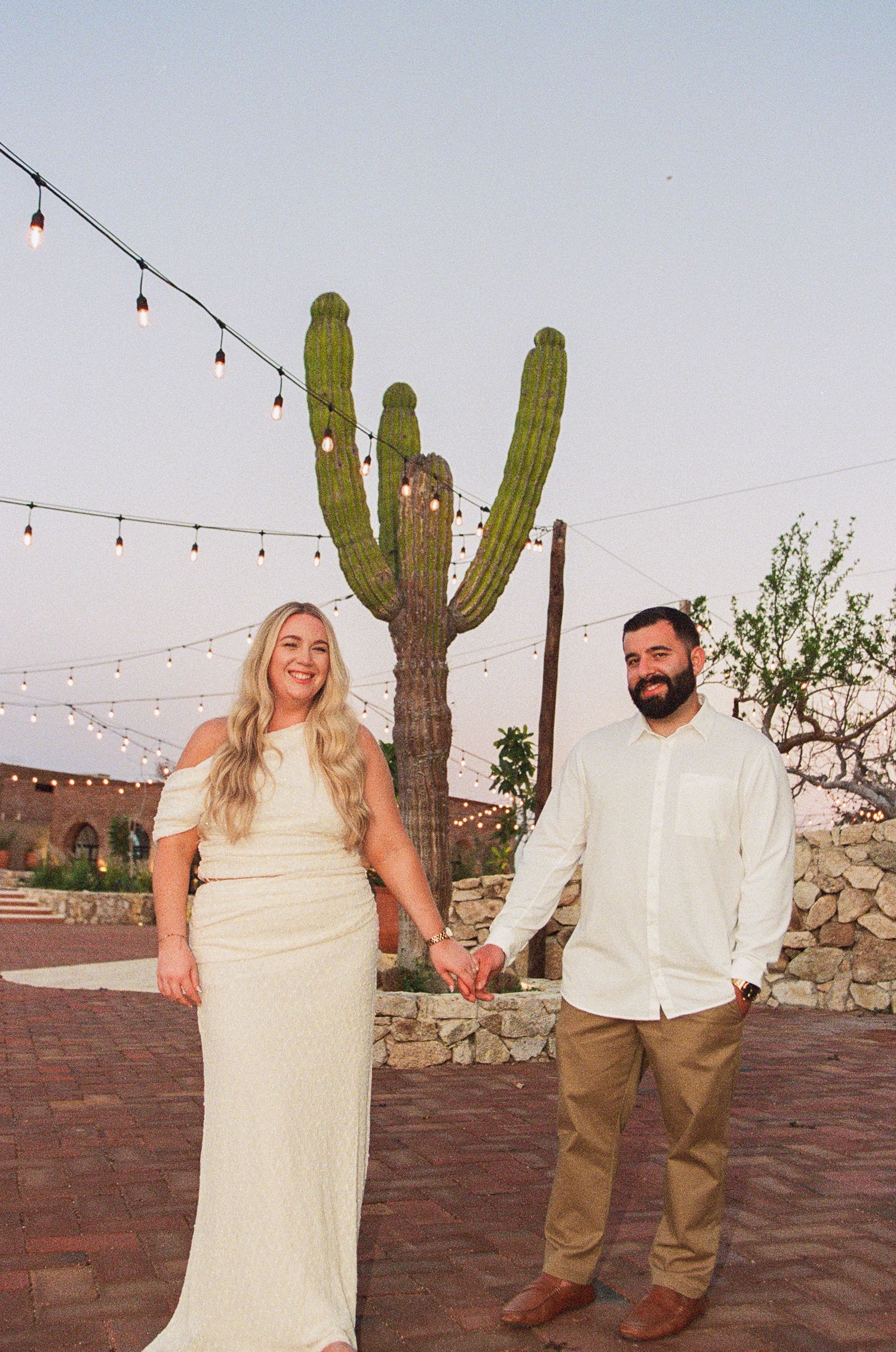 Engaged couple holding hands in front of a tall saguaro cactus with string lights at an outdoor desert venue Monte Cardon.