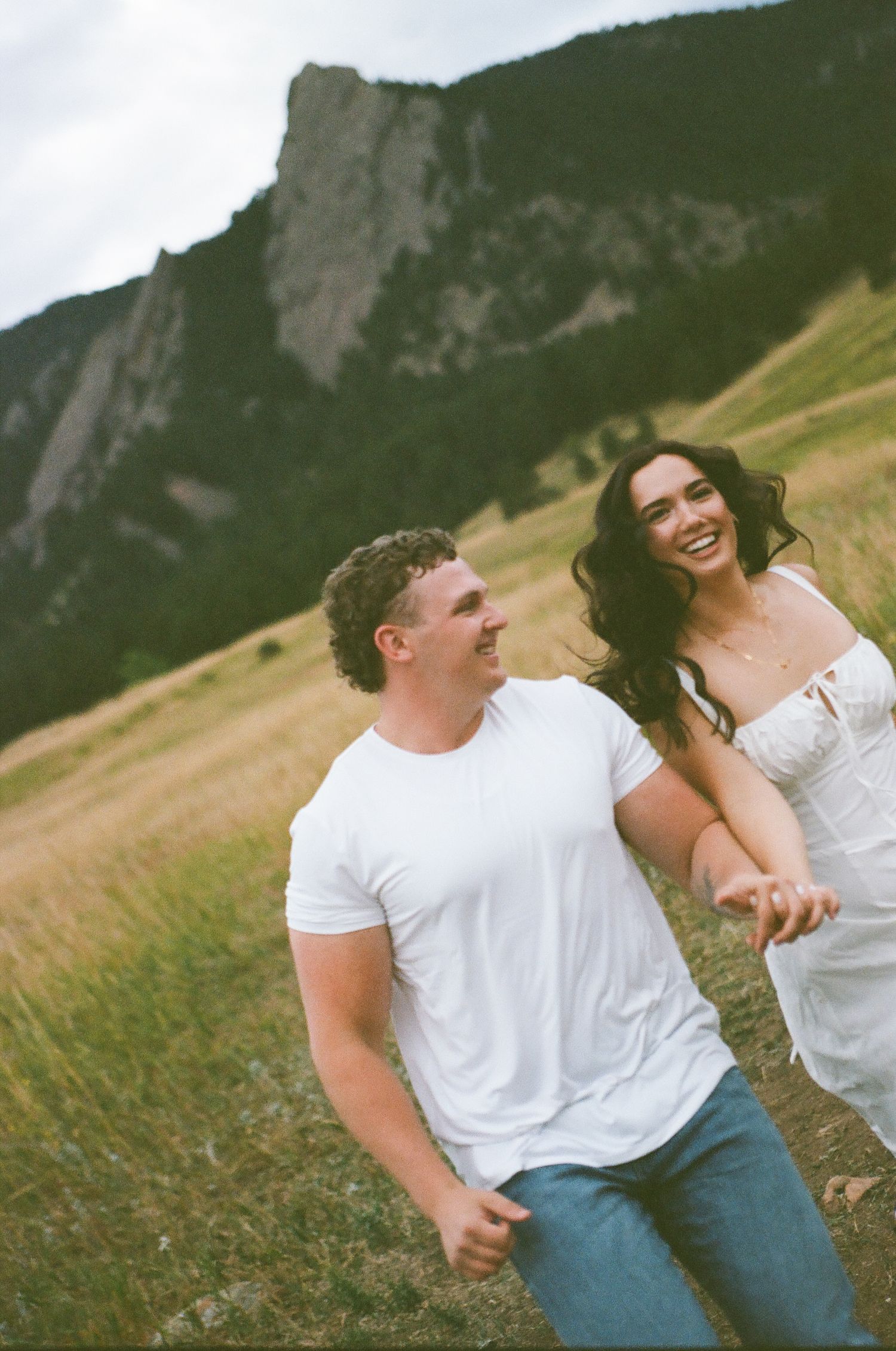 A smiling couple in white outfits holds hands in a grassy field with dramatic rocky mountain flatirons in the background.