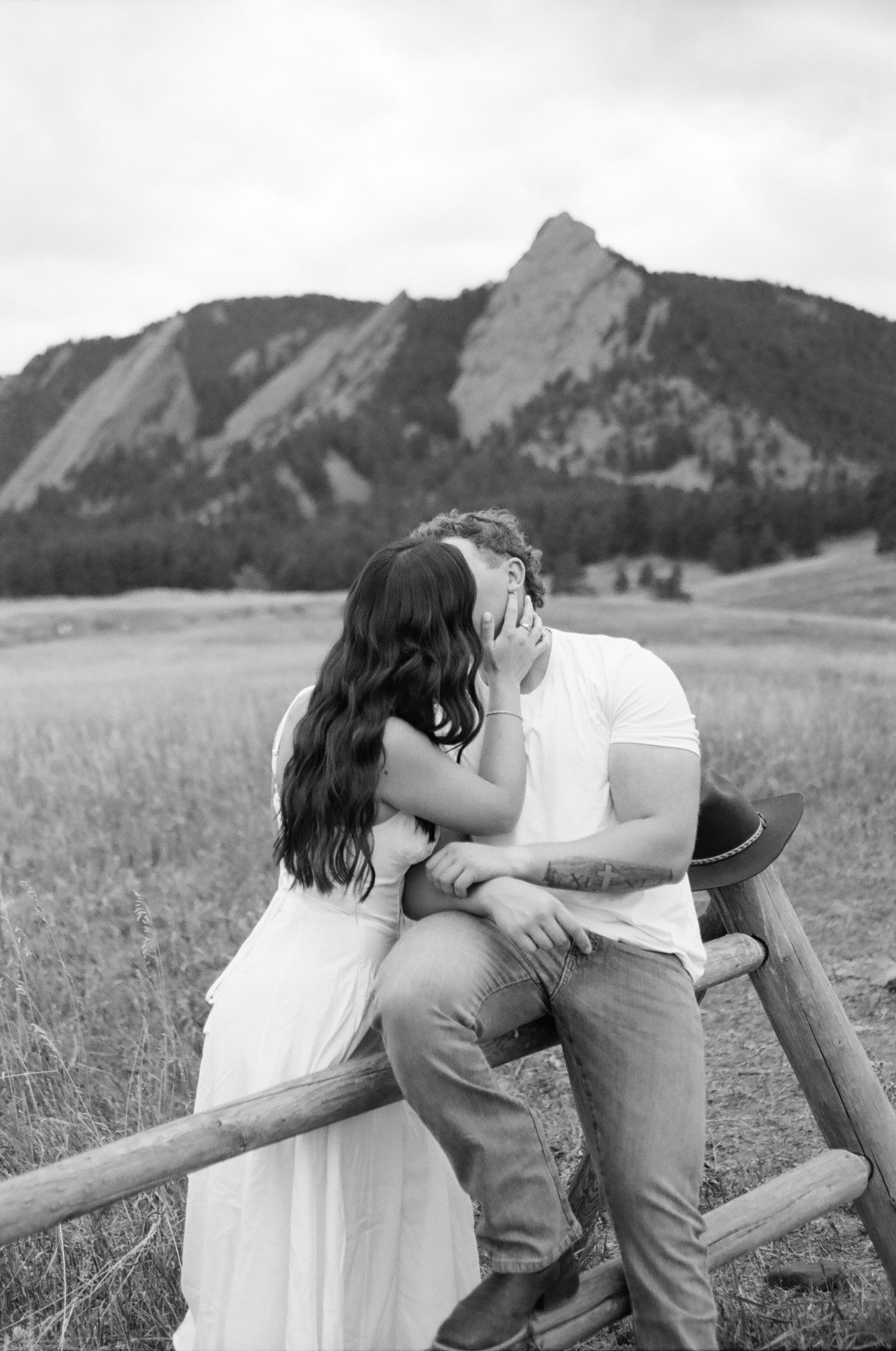 Black and white photo of a couple kissing by a wooden fence with the Flatirons mountains in Boulder, Colorado behind them.
