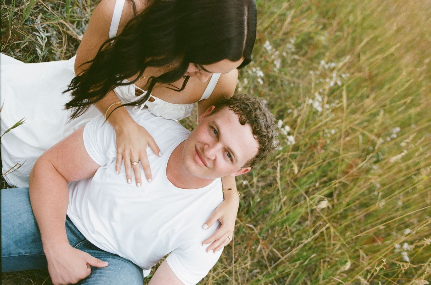 Editorial photo of couple in a grassy field, man lying down smiling up at woman leaning over him, outdoor engagement in Boulder Colorado.