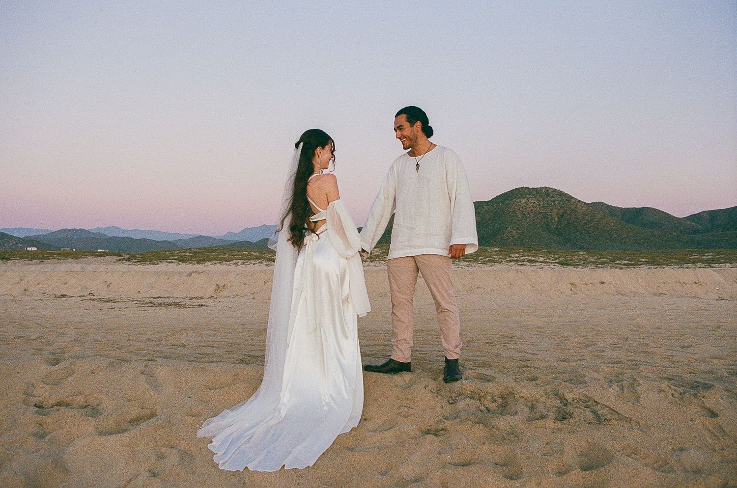 Bride and groom hold hands on sandy beach at sunset with mountains in background during outdoor wedding ceremony near Todos Santos, Mexico.