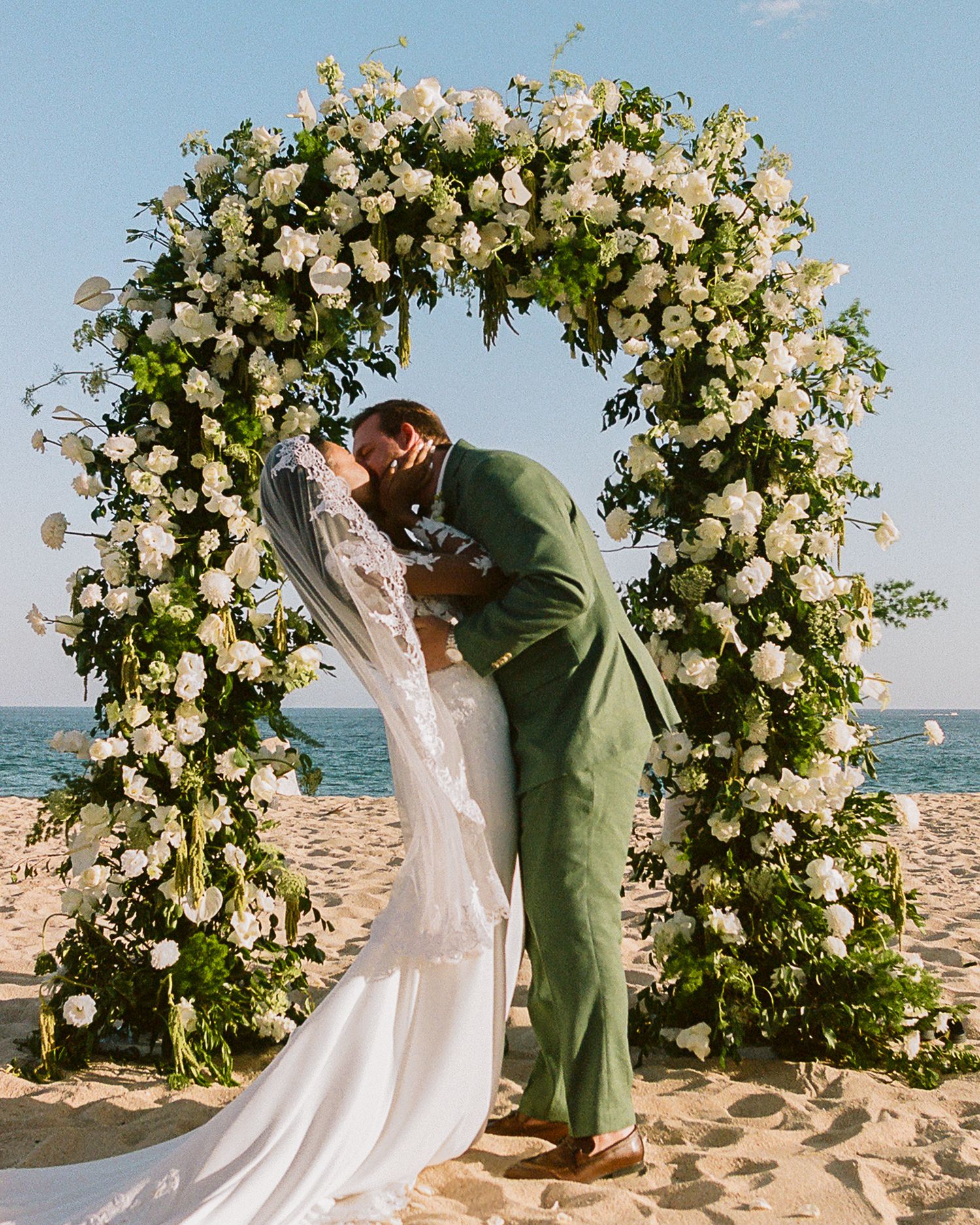 Couple shares first kiss under white floral arch on sandy beach at outdoor wedding ceremony in Cabo San Lucas, Mexico.