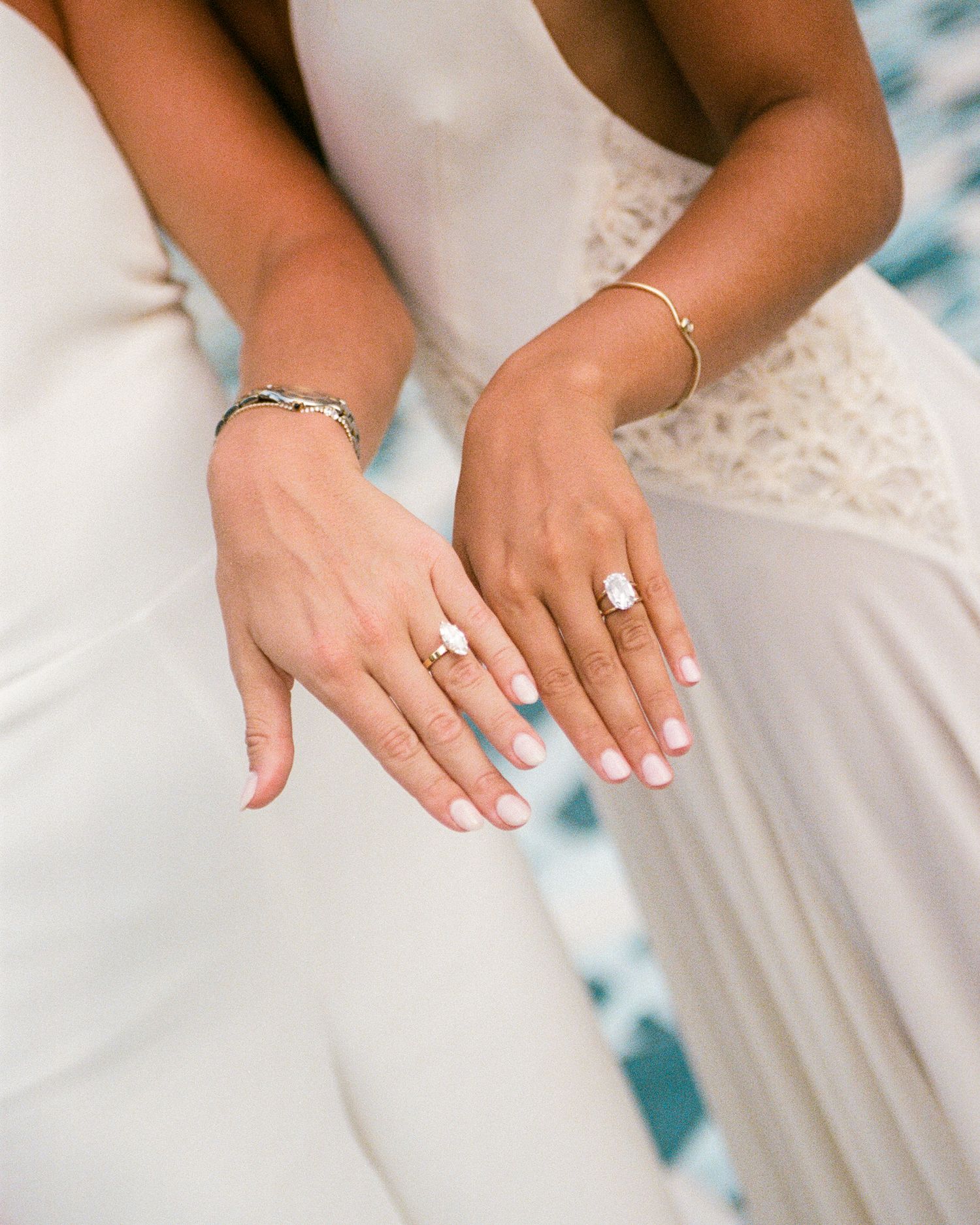 Two brides in white gowns display their diamond engagement rings and gold bracelets side by side.