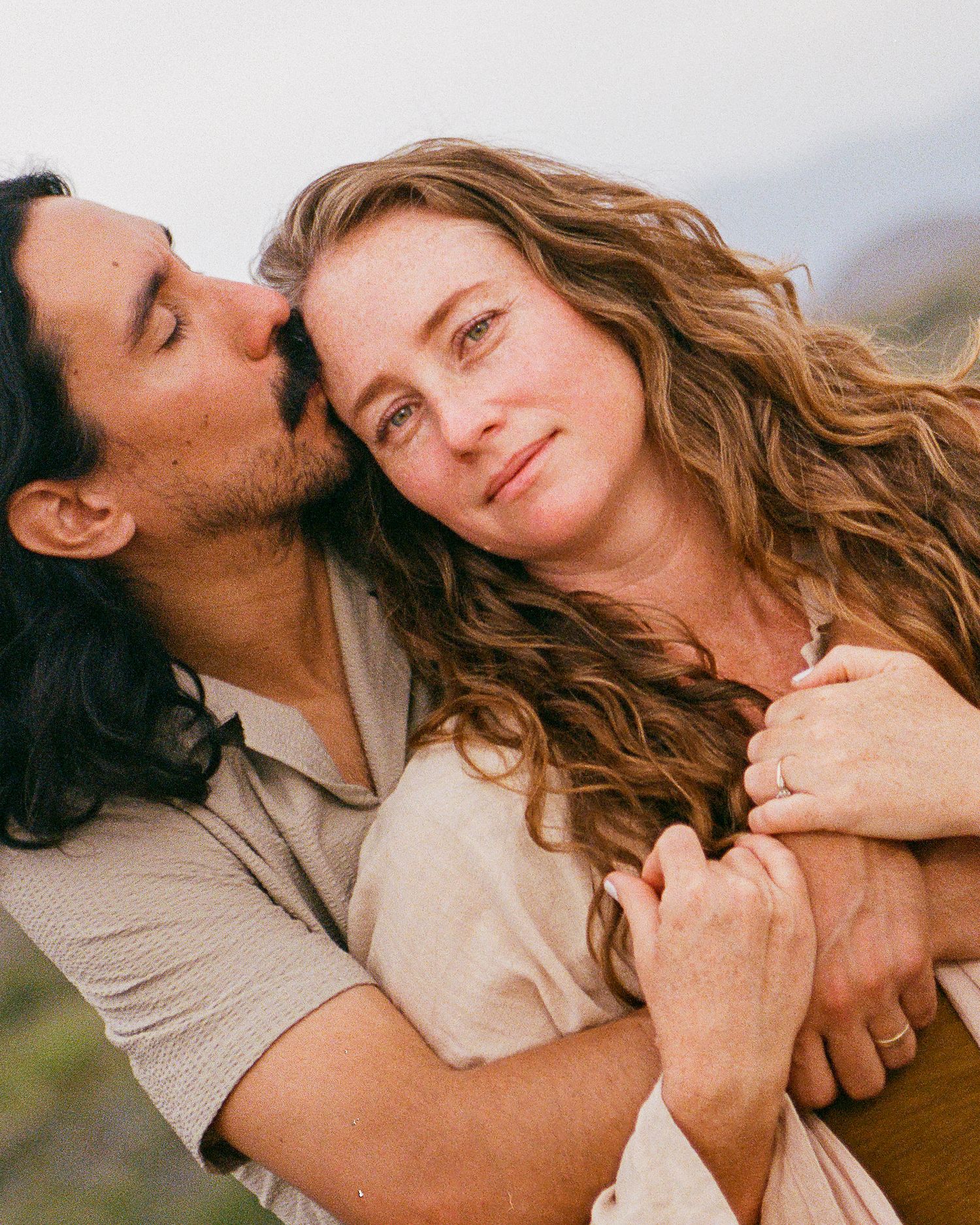 A couple shares a tender embrace outdoors, man kissing woman's cheek while she smiles softly, wearing casual earth-toned clothing.