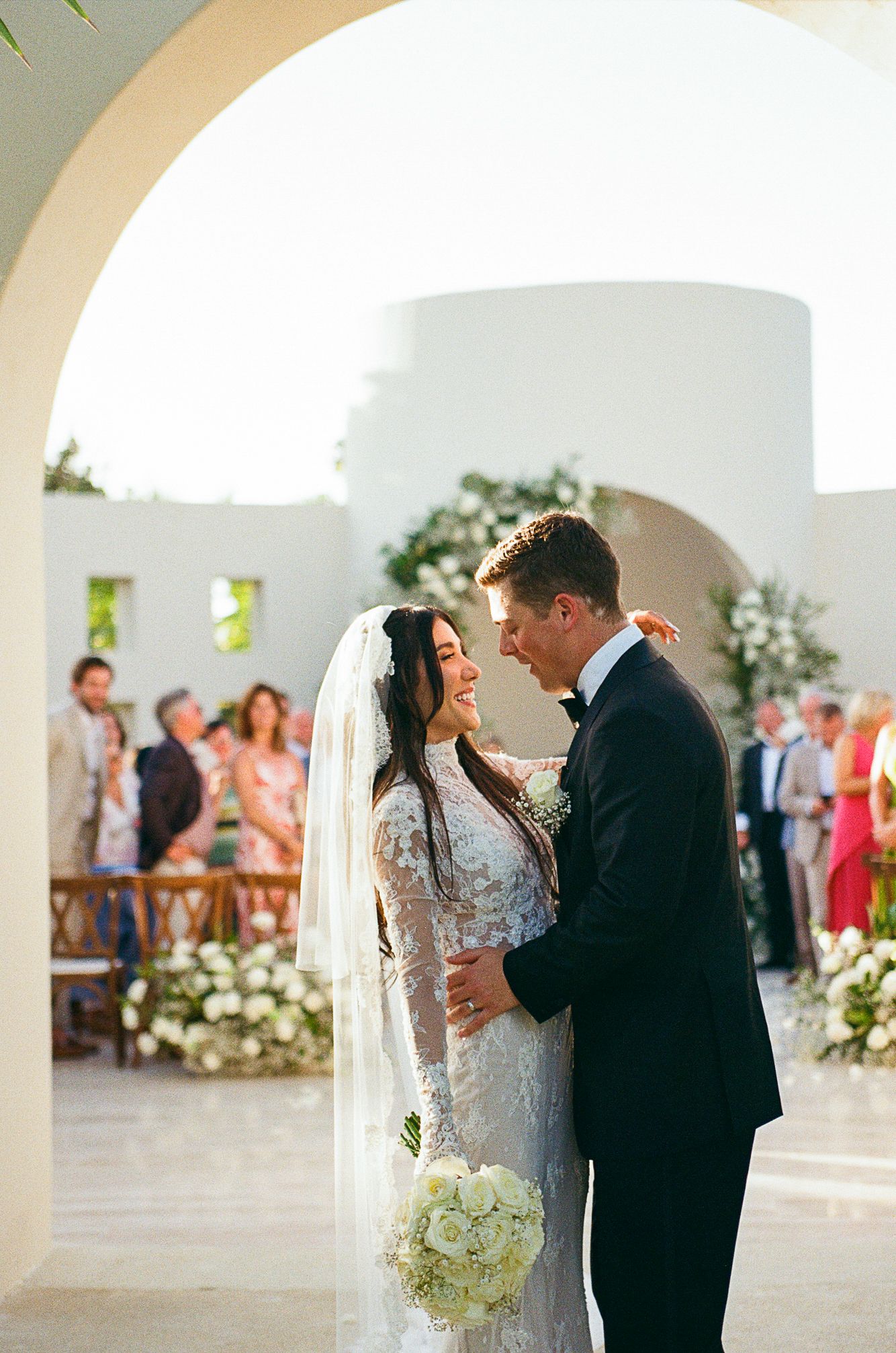 Bride and groom share a romantic moment at an outdoor wedding ceremony with white floral arrangements and guests seated behind at Baja Luna.