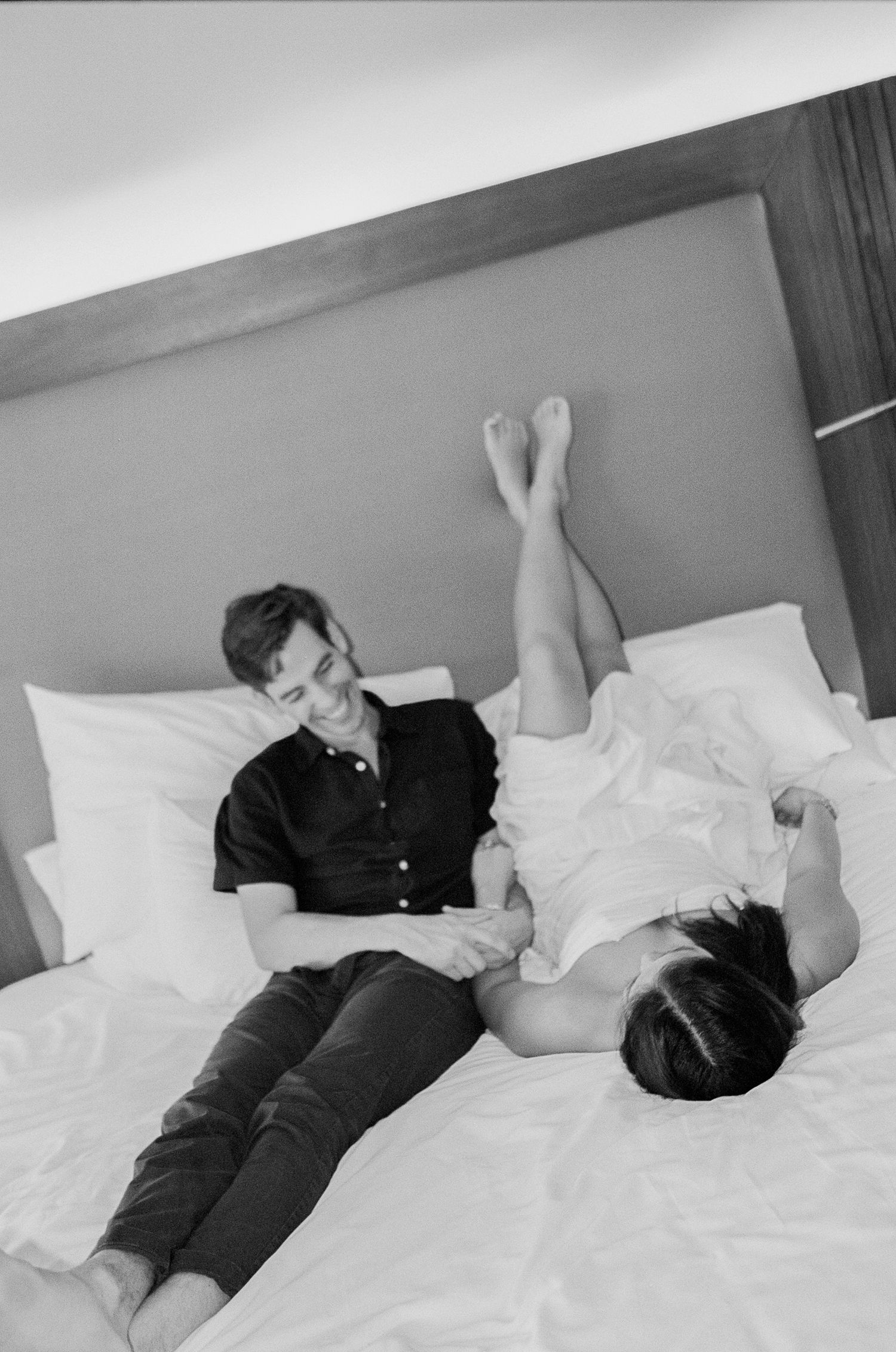 Black and white photo of a couple relaxing on a white hotel bed, smiling and enjoying each other's company during engagement session.