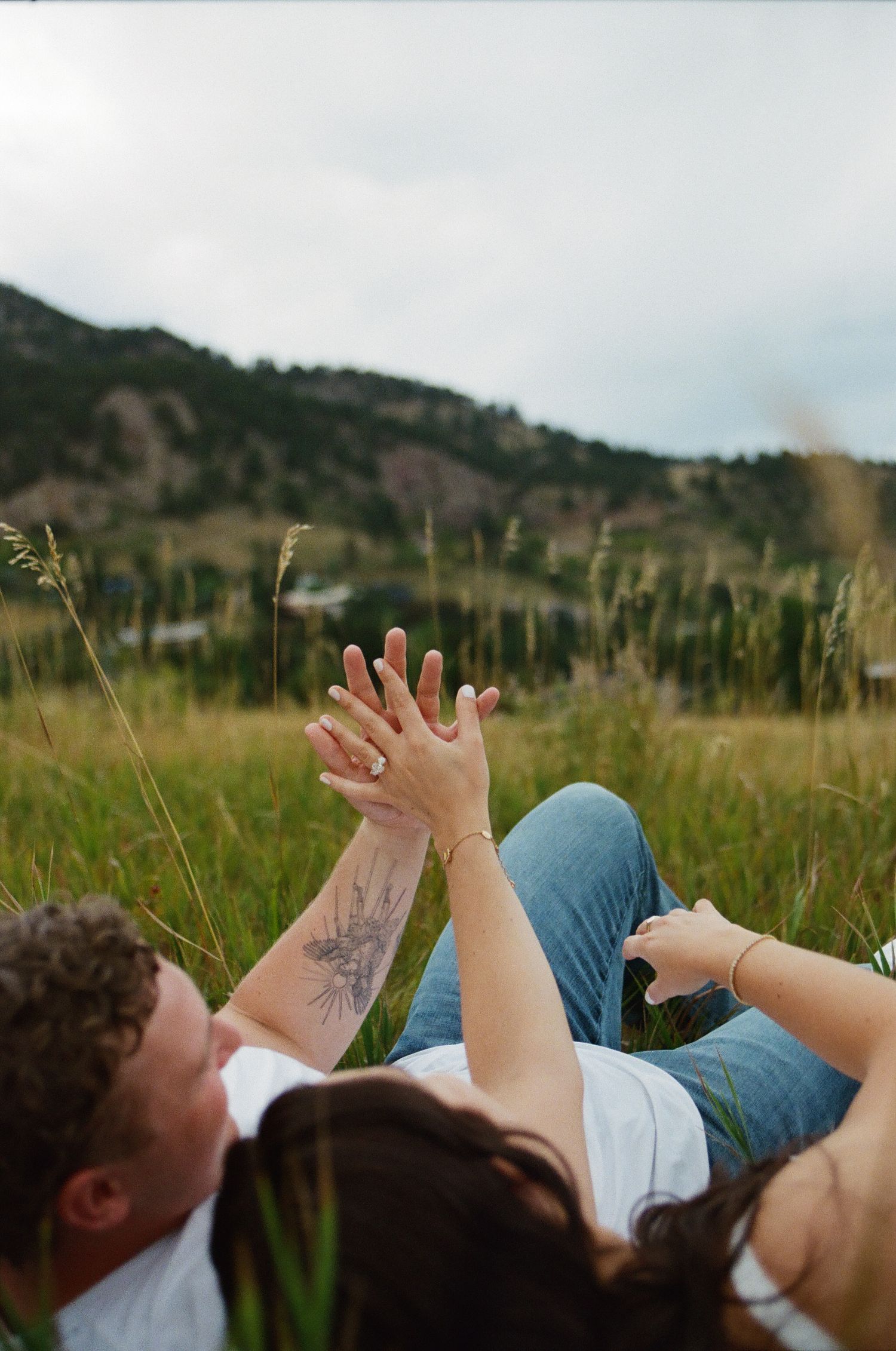 Couple lying in a grassy mountain meadow, holding hands up showing engagement ring, with tattoos visible in Boulder Colorado.