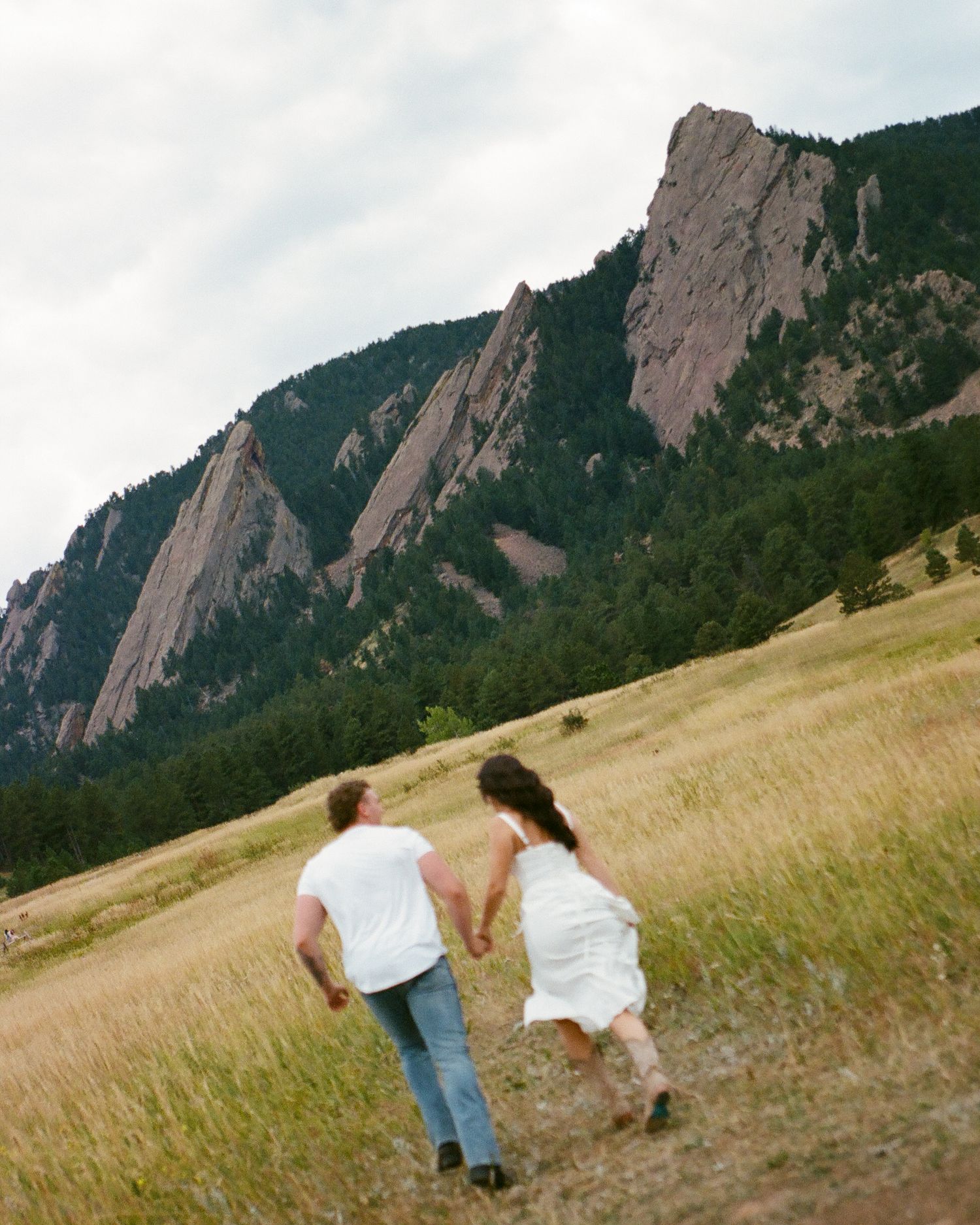 Couple in white holding hands running through golden meadow with dramatic Flatirons rock formations in Boulder, Colorado.