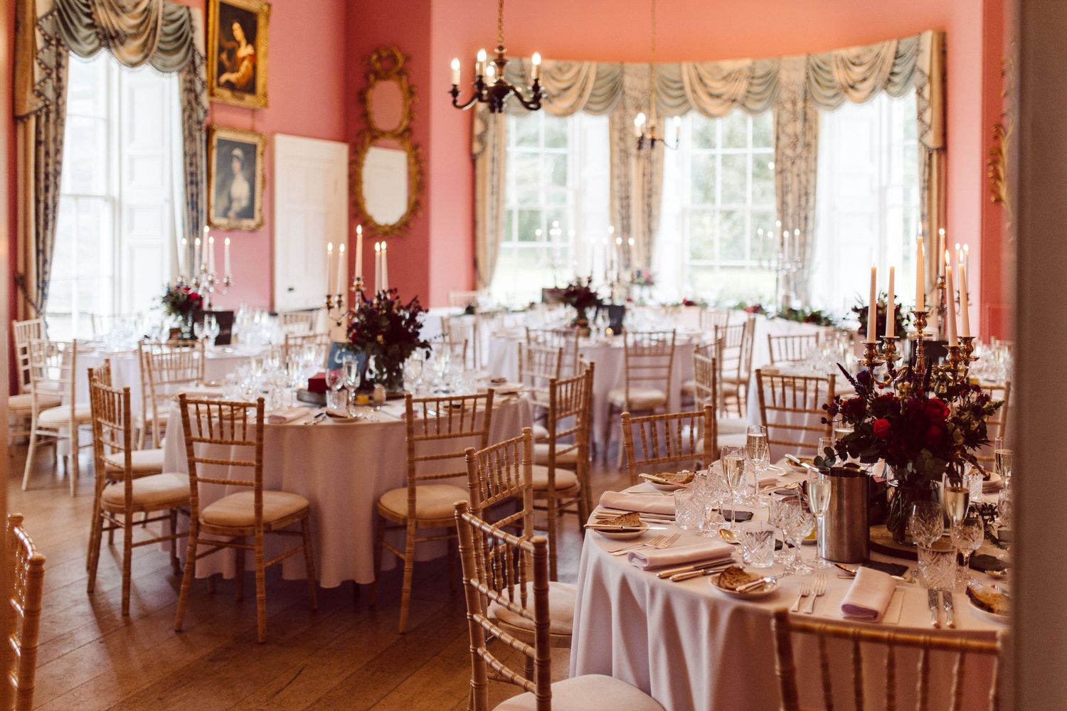 An elegant pink ballroom with white tables, gold chairs, and crystal chandeliers set up for a formal reception.