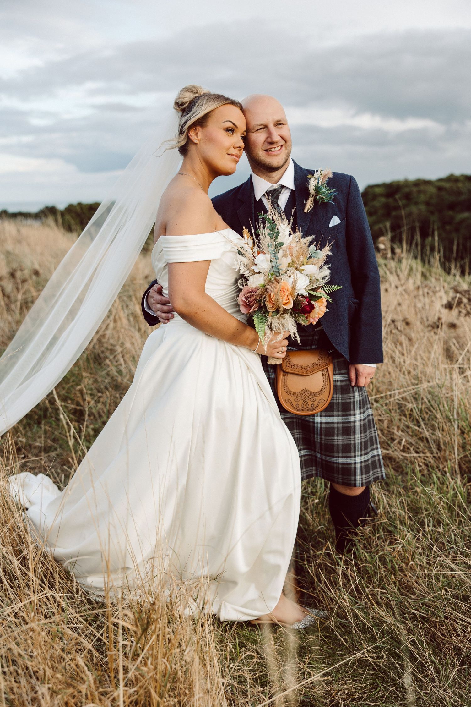 A romantic wedding couple embraces in a field of tall grass with flowing white veil and Scottish tartan details.
