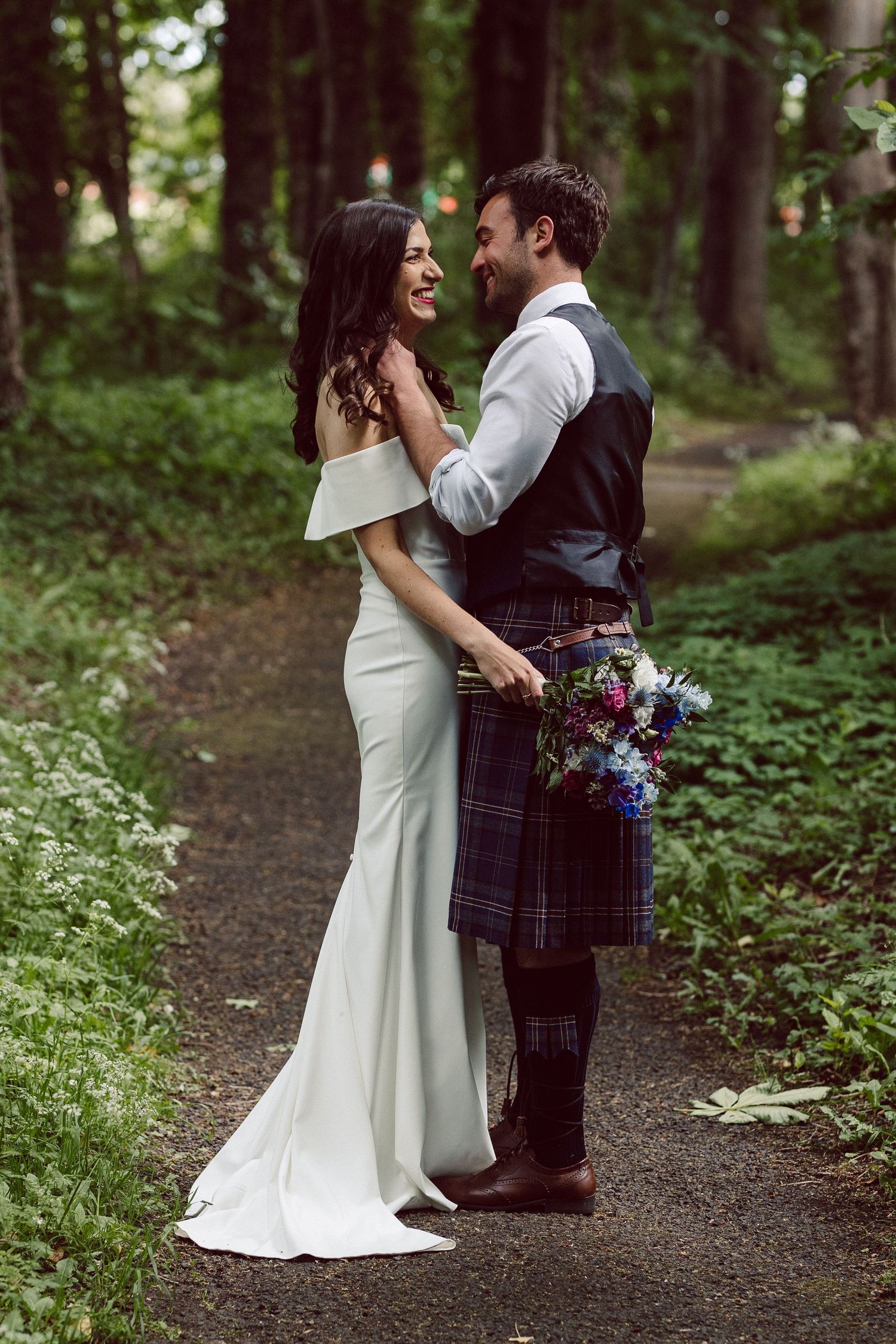 A couple shares an intimate moment on a wooded forest path during their Scottish-themed wedding celebration.