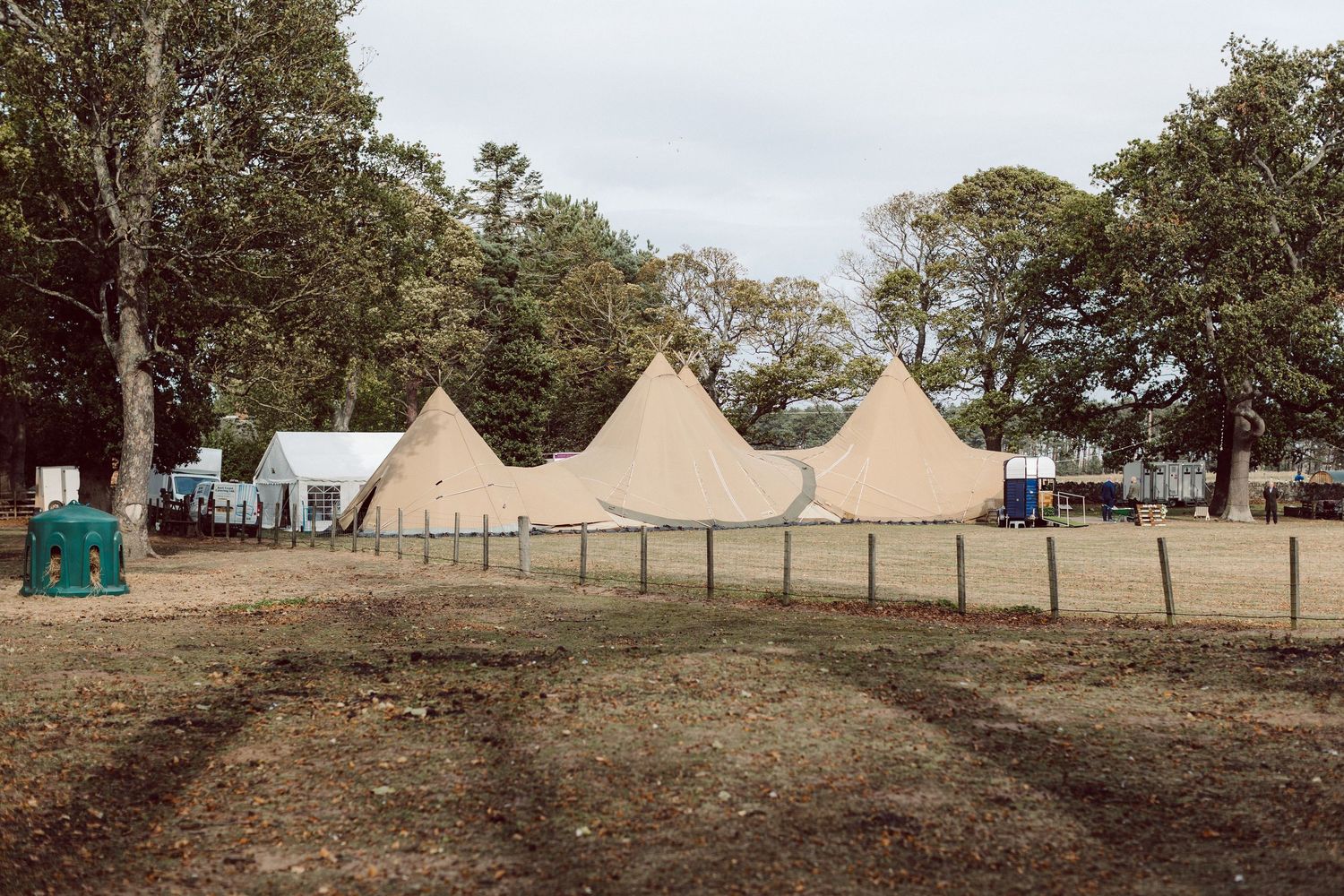 A panoramic view of rustic camping tents set up in a grassy field surrounded by trees on a cloudy day.
