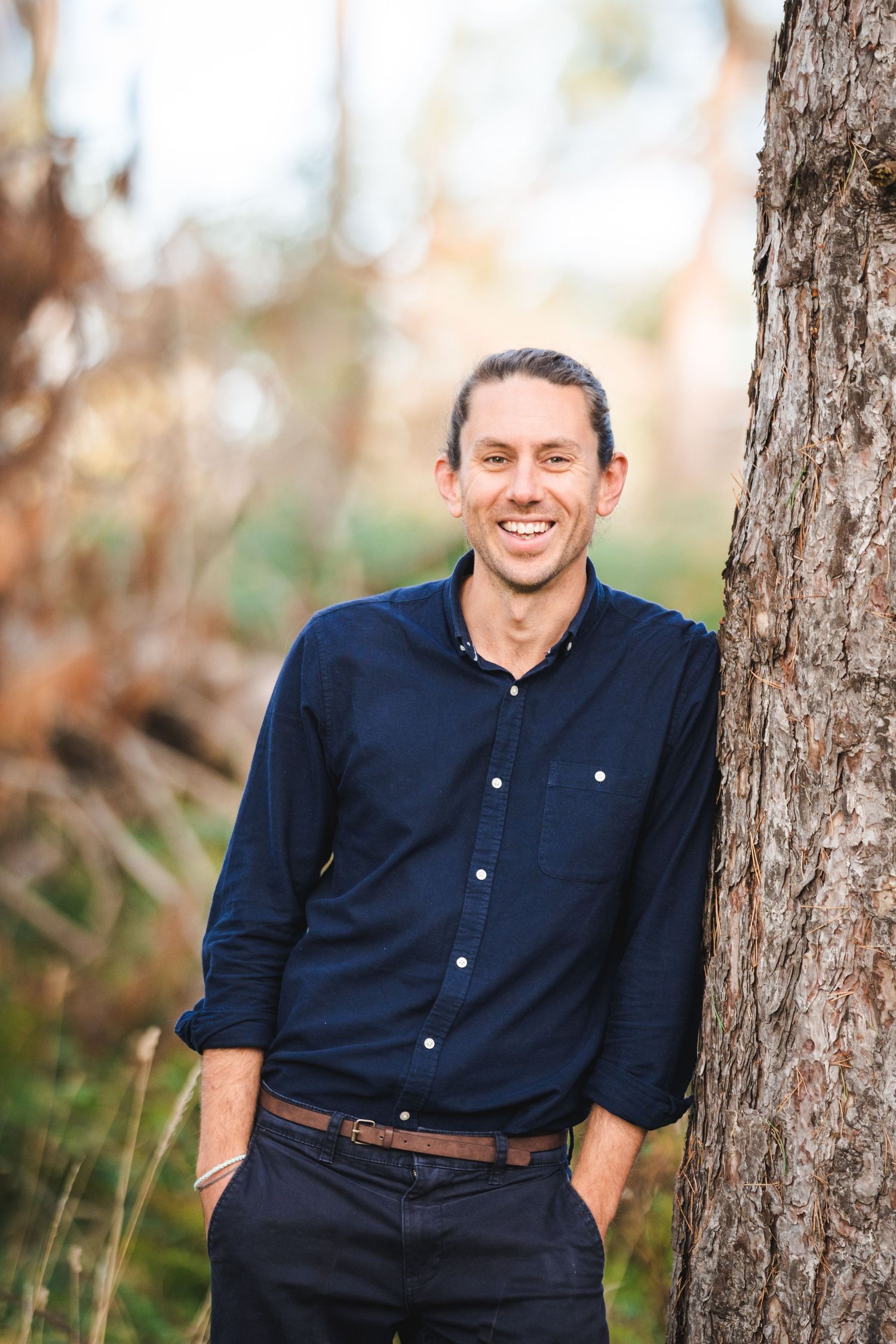 A person in a navy button-down shirt and dark pants stands casually leaning against a tree trunk outdoors.