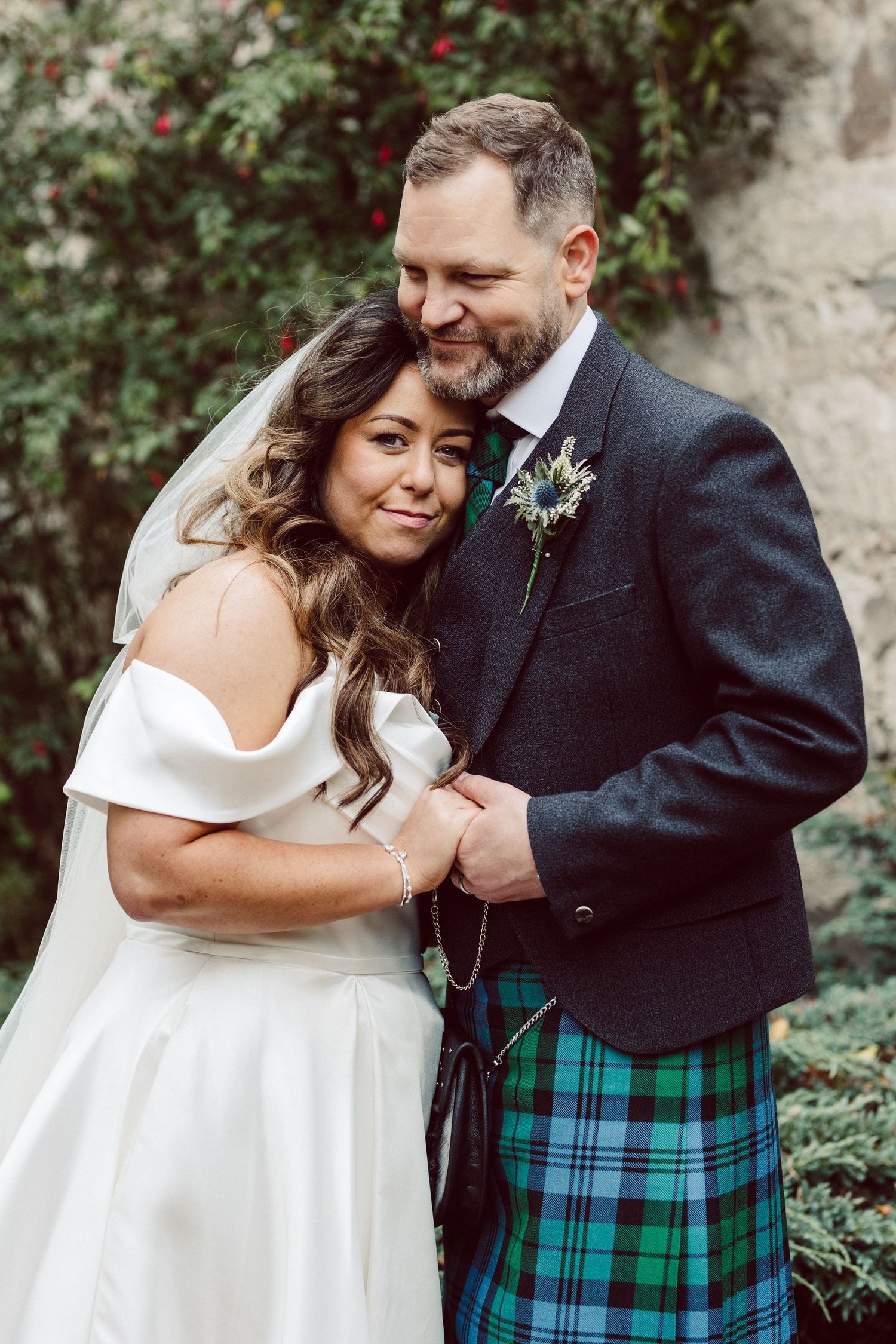 A couple shares joyful moments in a garden setting, with the groom wearing a traditional Scottish kilt and black jacket.