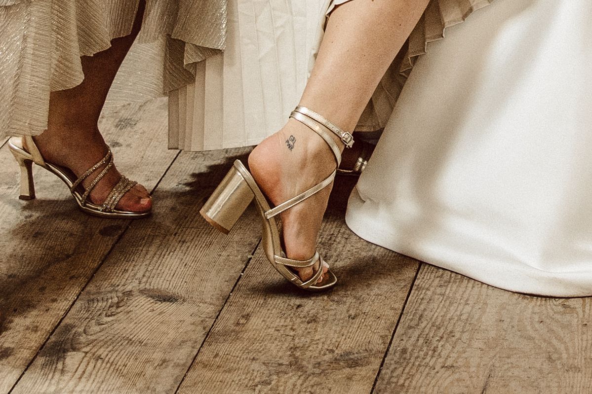 Gold block heel sandals with ankle straps shown on wooden floor next to flowing white dress fabric.