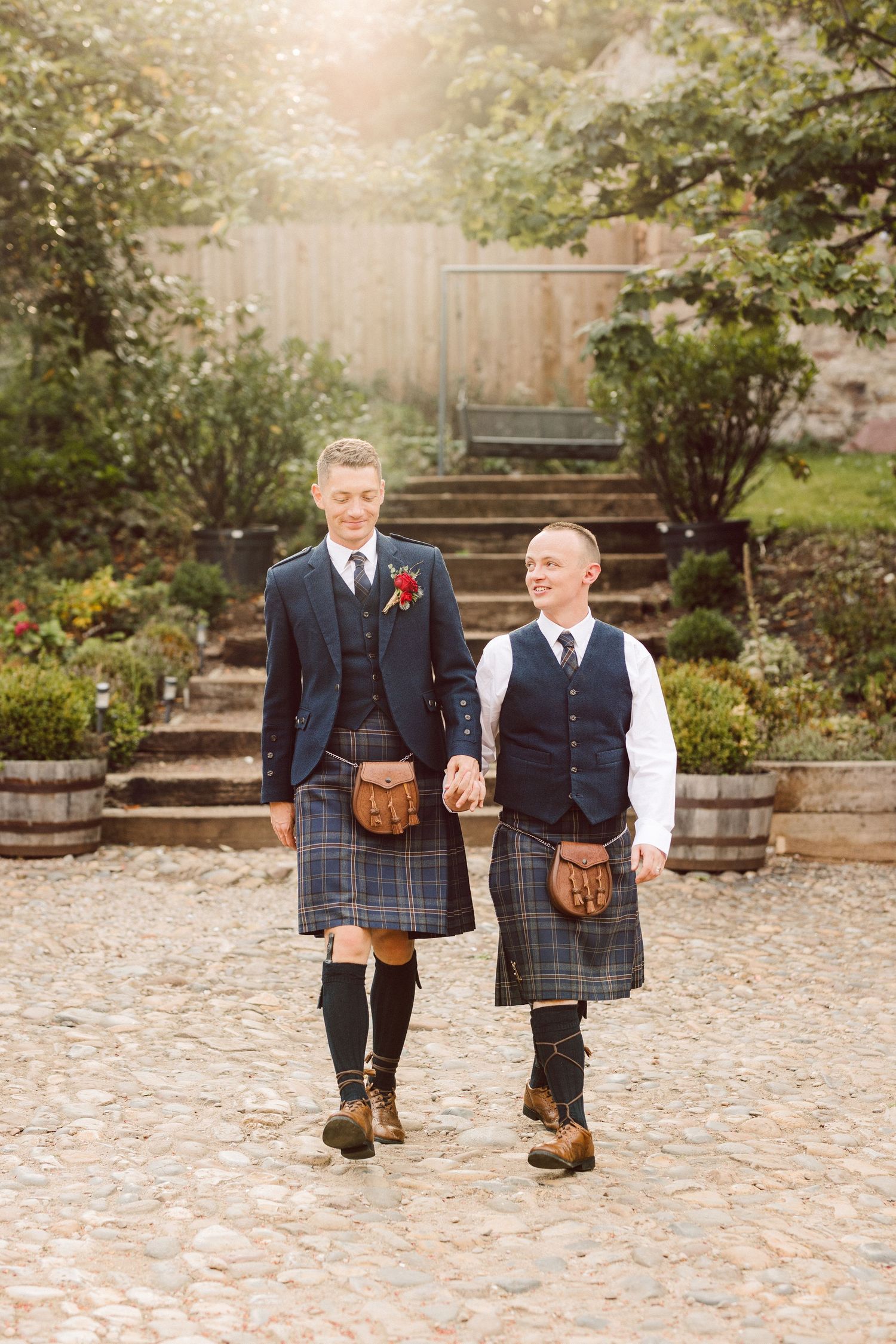Two people in traditional Scottish kilts and formal jackets walk together on cobblestones in a garden setting.