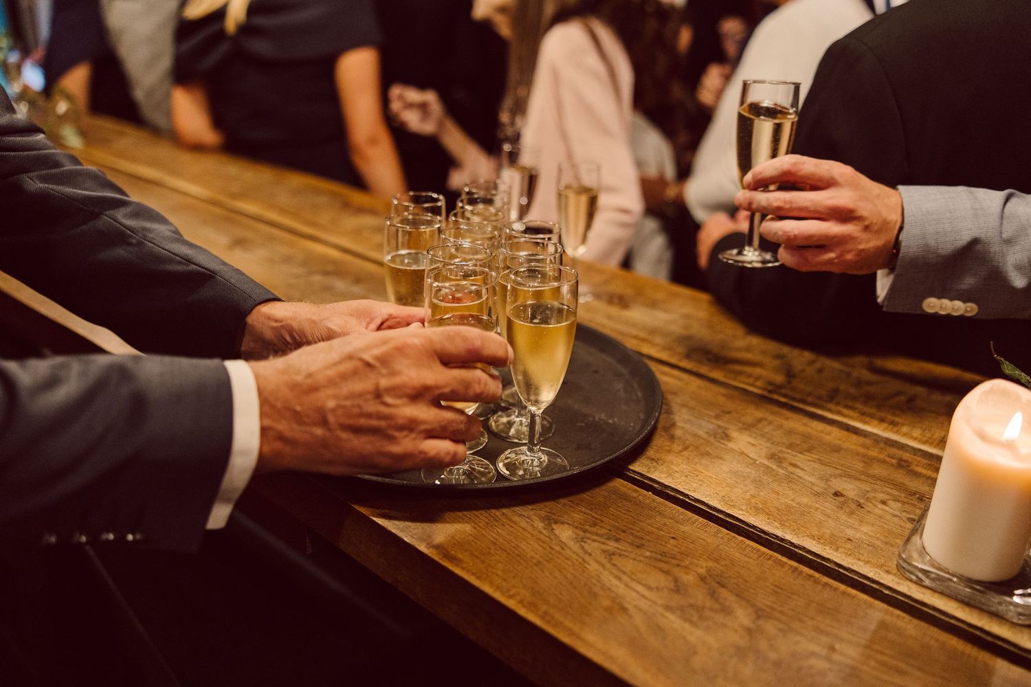 Hands holding champagne glasses for a toast at a wooden bar with candlelight.