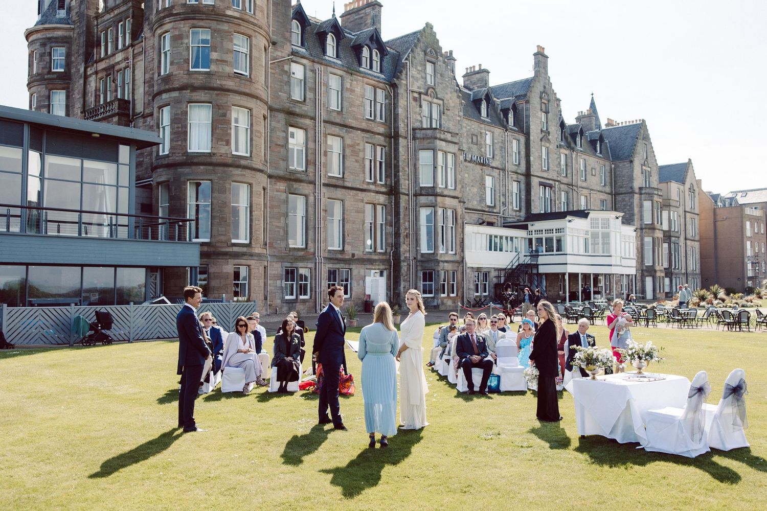 Panoramic sequence of Victorian buildings along a beachfront promenade with people walking and enjoying the sunny day.