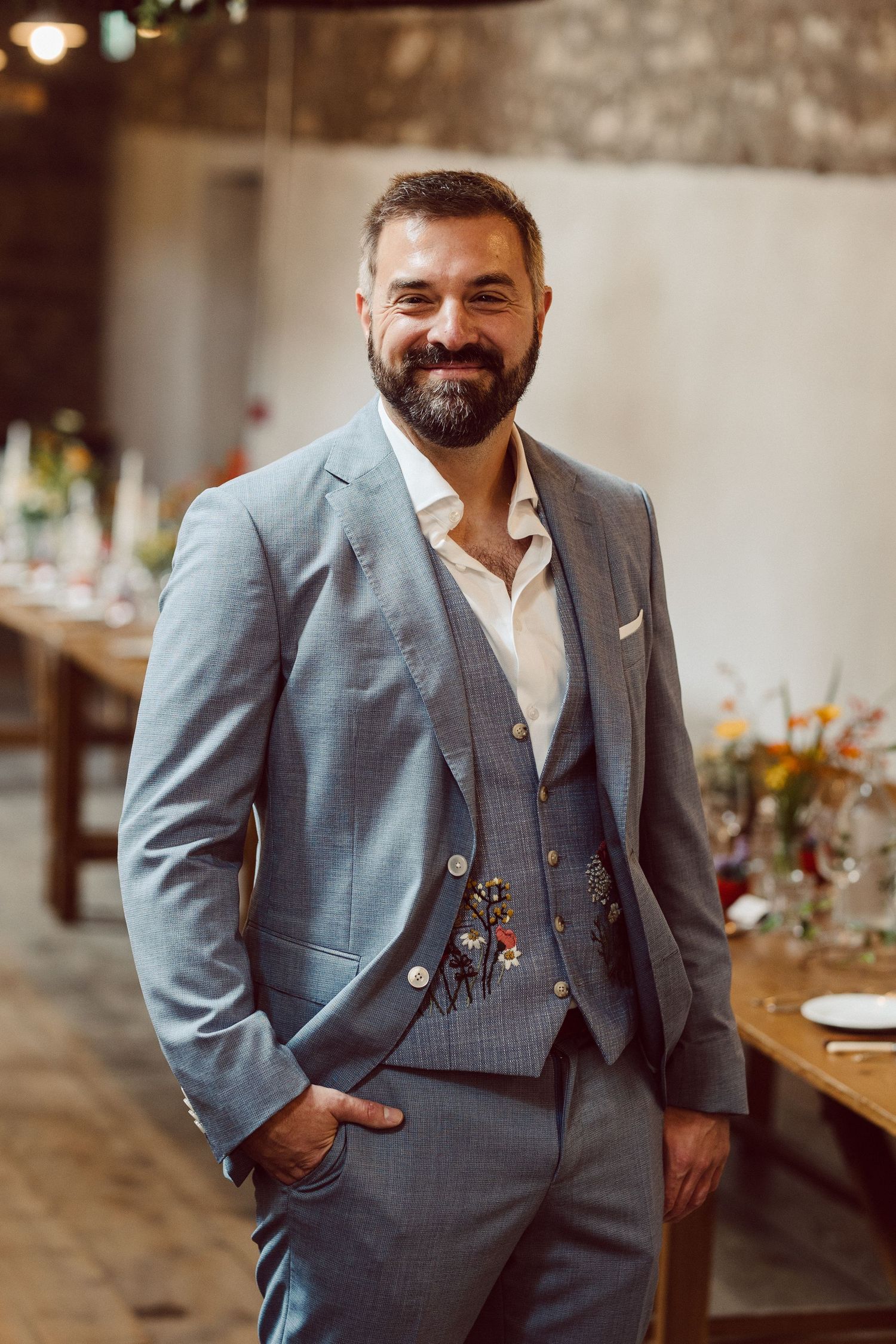 A bearded person wearing a light blue three-piece suit with a white shirt smiles warmly in an indoor venue setting.