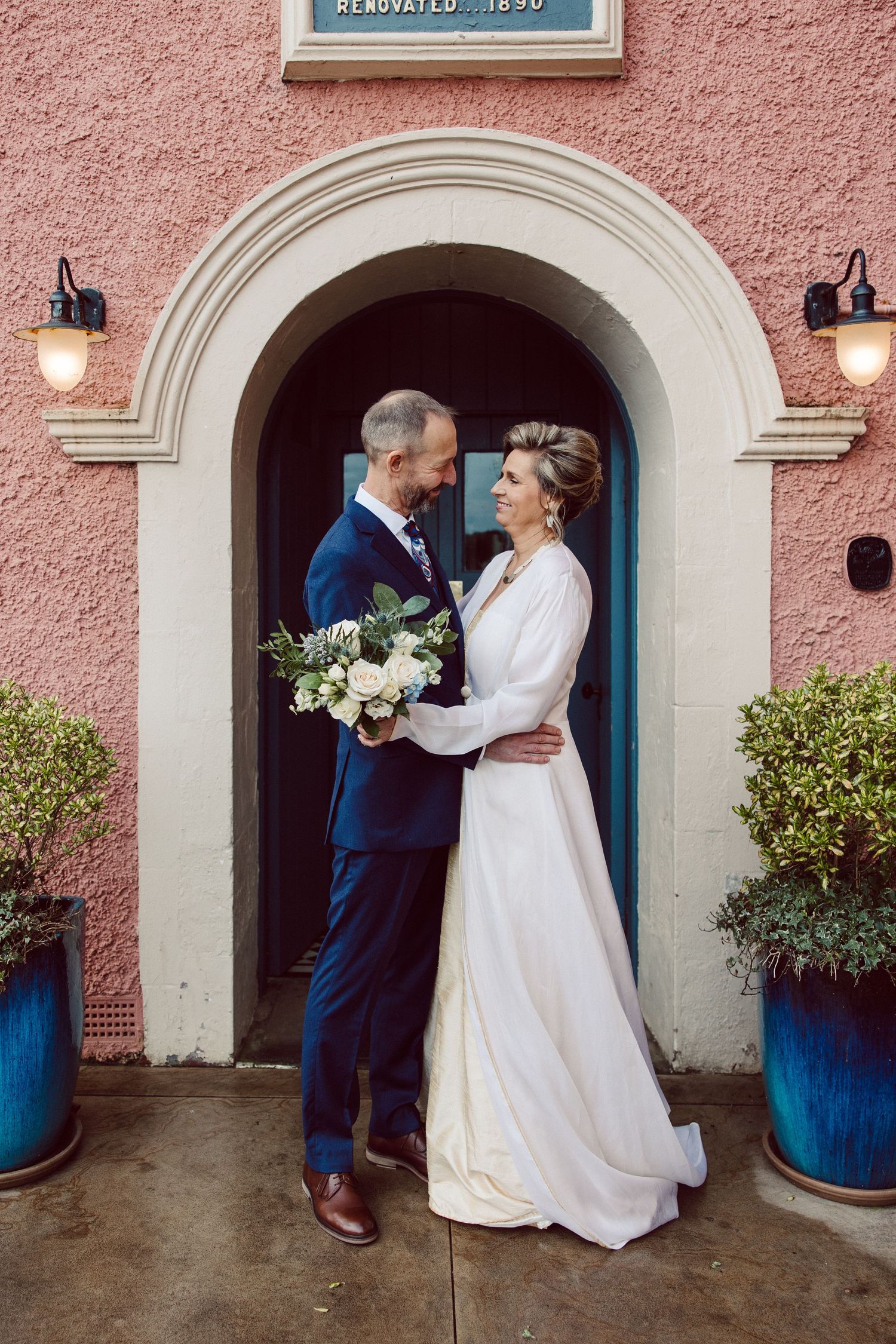 A wedding couple shares romantic moments in front of a pink stucco building with arched doorways and blue planters.