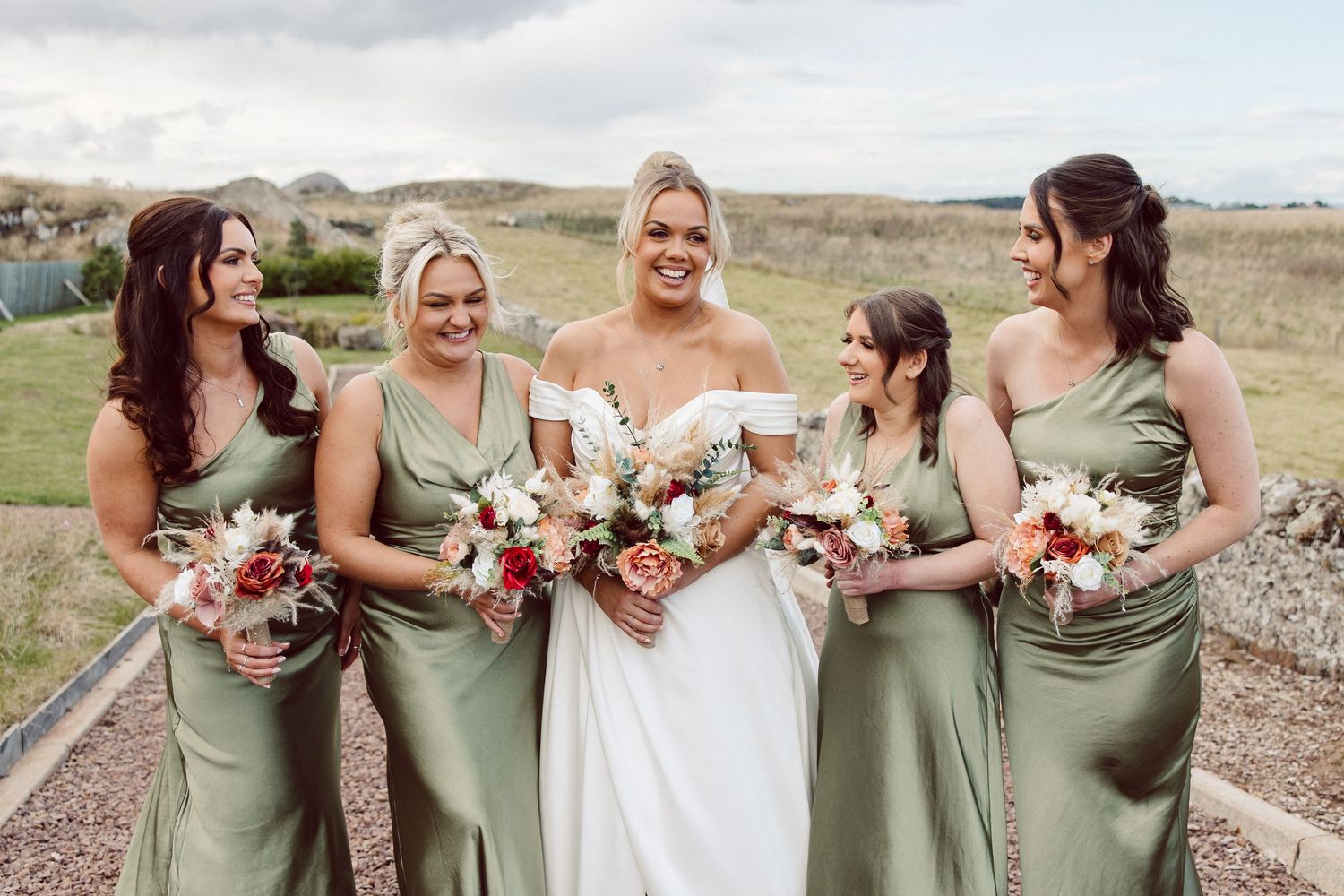Wedding party in sage green dresses poses outdoors holding pink and white floral bouquets on a sunny day.