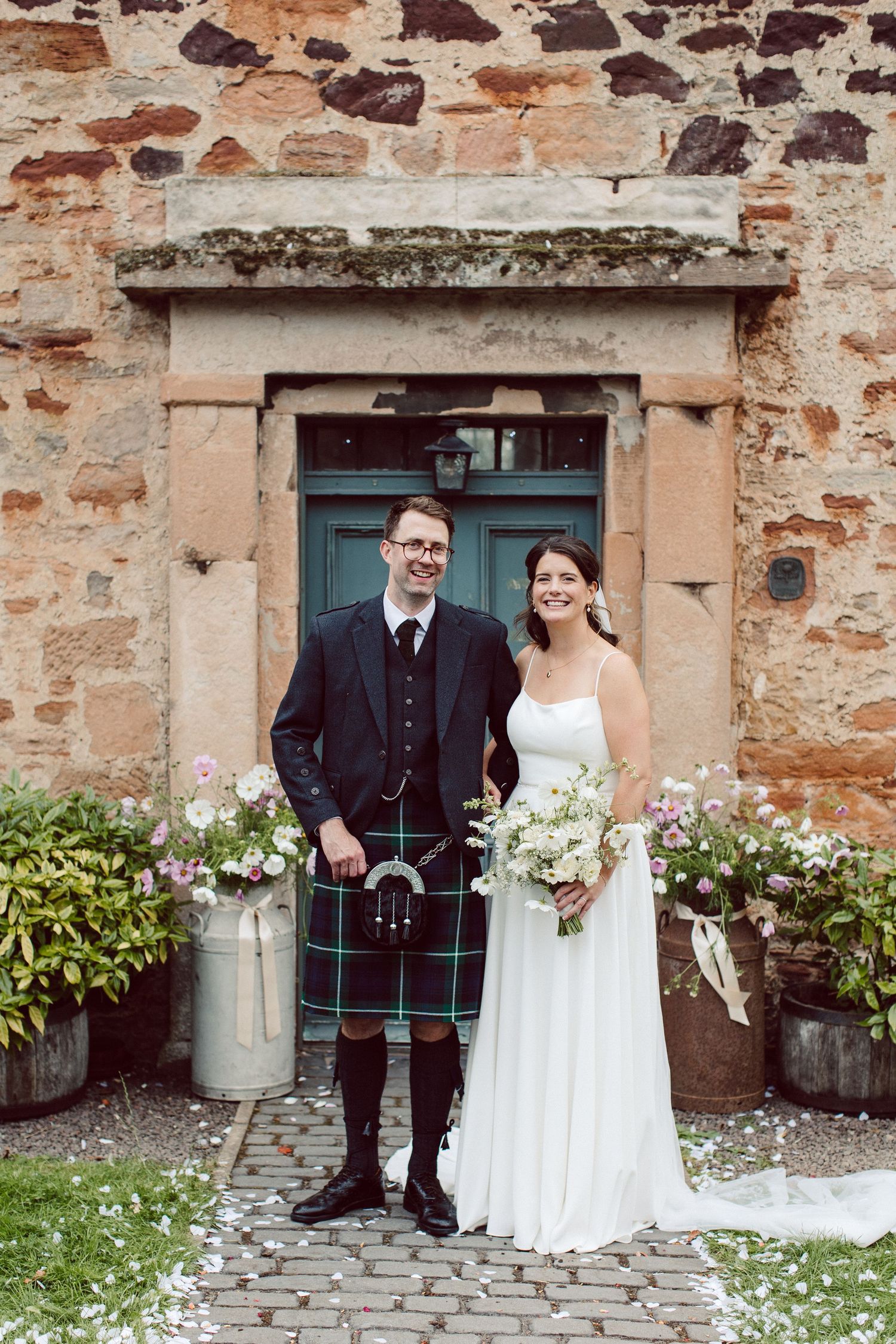 A traditional Scottish wedding couple stands in front of a rustic stone wall and blue door with flower decorations.