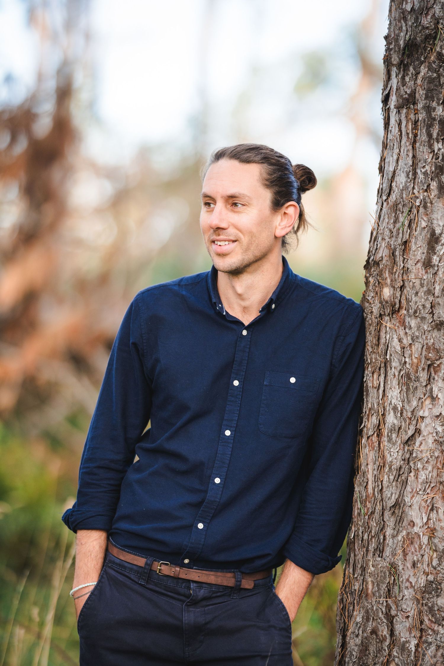A person wearing a navy blue button-down shirt poses casually against a tree trunk in an outdoor setting.