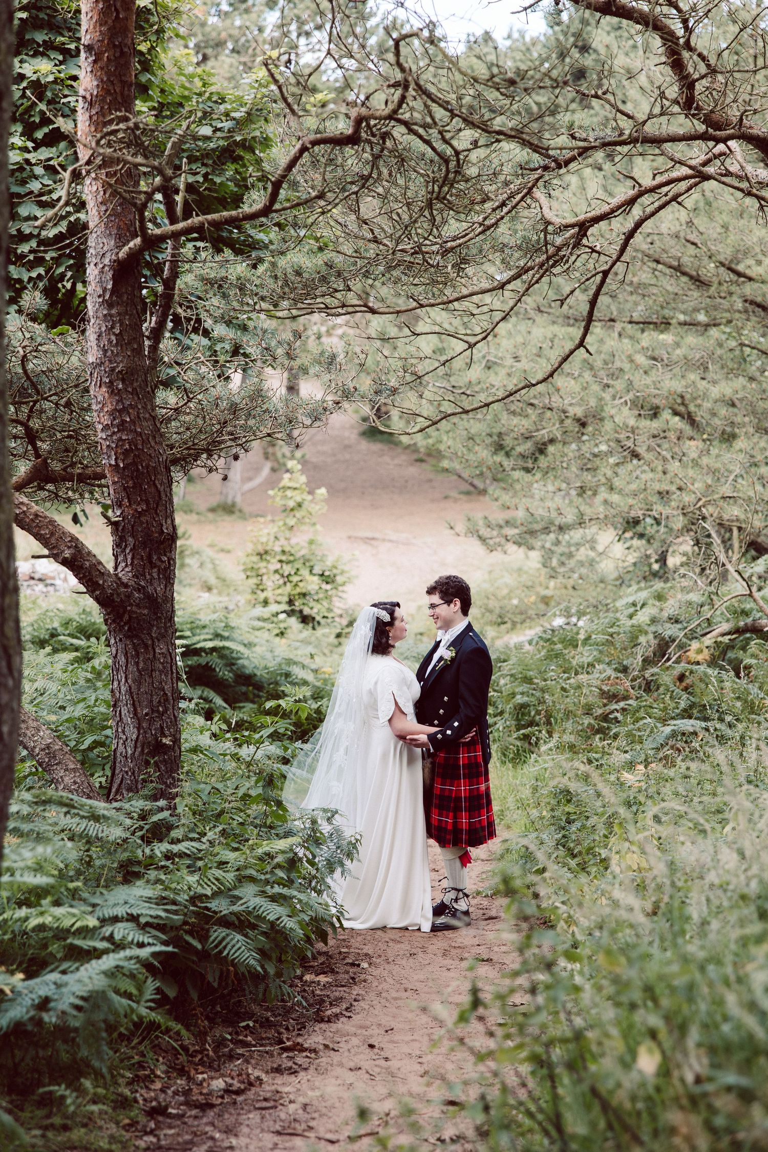 A couple walks along a serene forest path surrounded by tall pine trees and lush greenery.