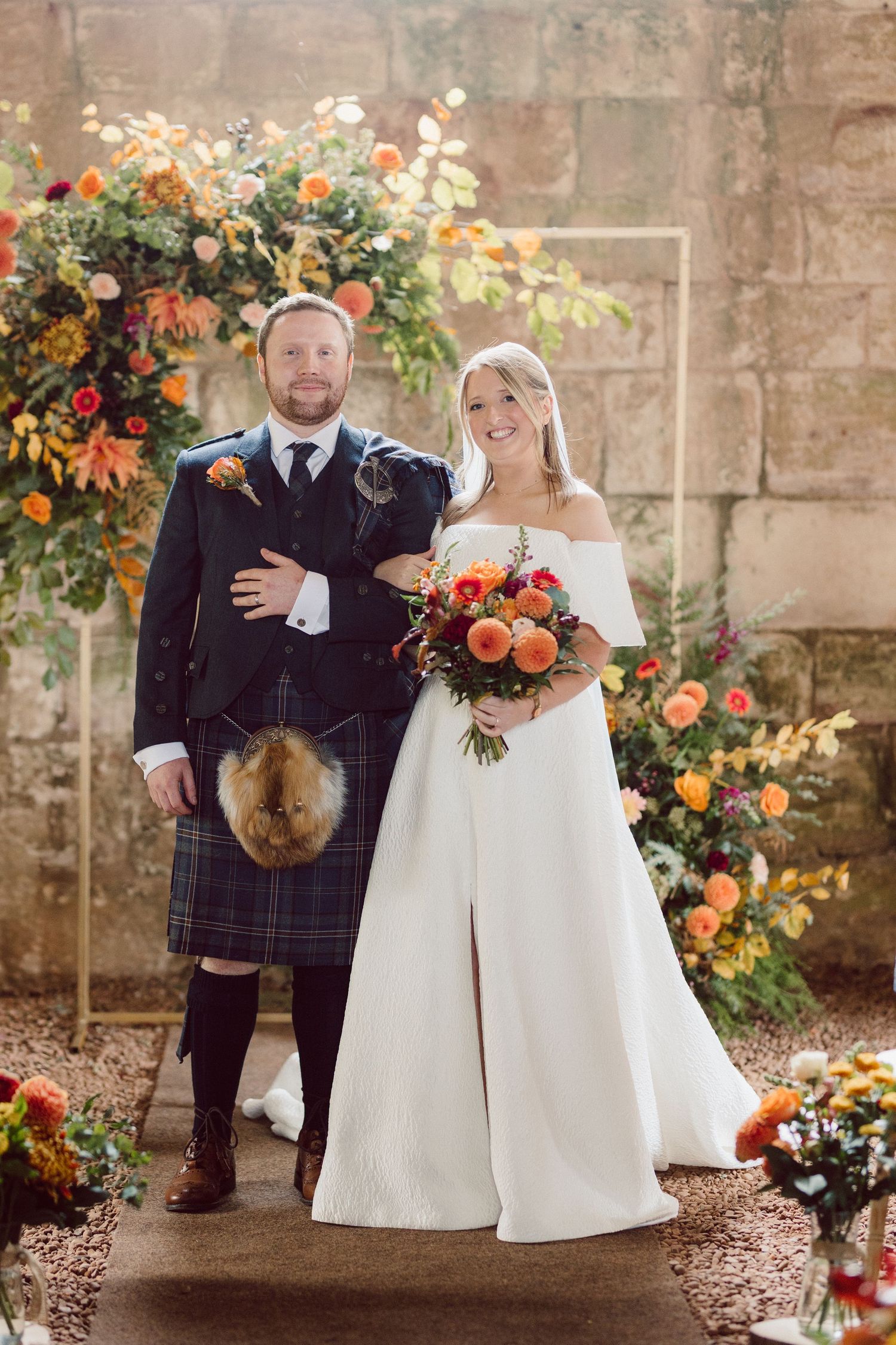 A wedding couple poses in traditional Scottish attire with orange and white floral arrangements in a stone courtyard.