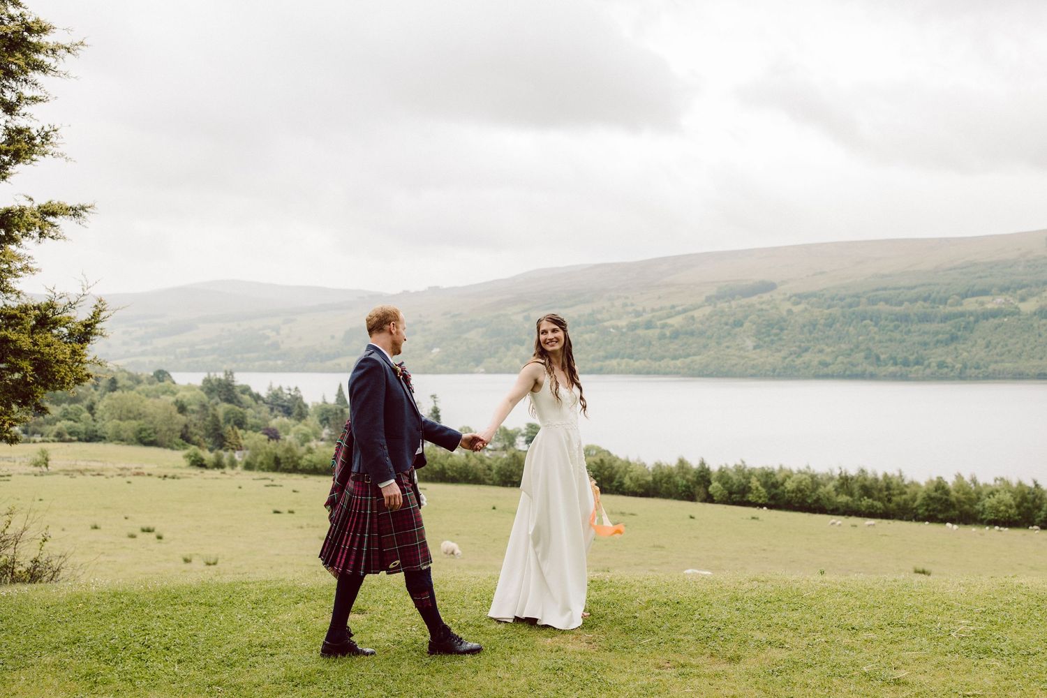 A couple in formal attire walks through a scenic hillside meadow overlooking a misty mountain landscape.