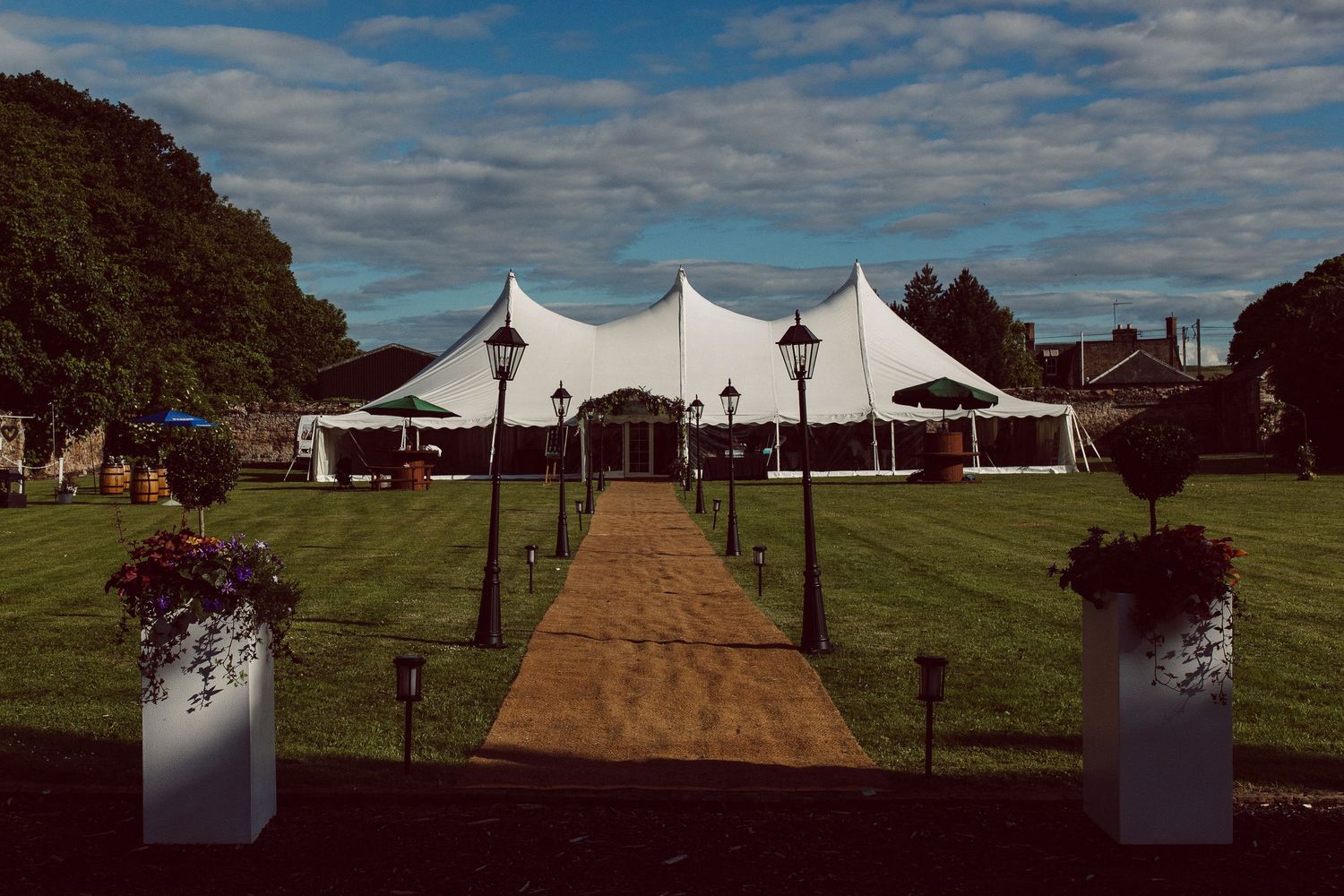 A series of white wedding tents set up in a grassy field with a path leading to the entrance under blue skies.