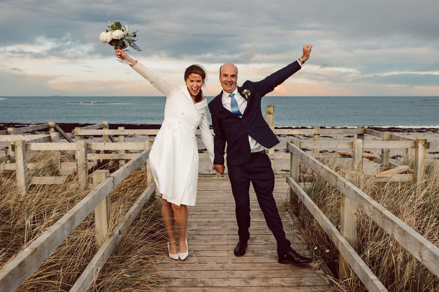 A joyful couple celebrates on a wooden boardwalk by the ocean with tall grass and cloudy skies in the background.