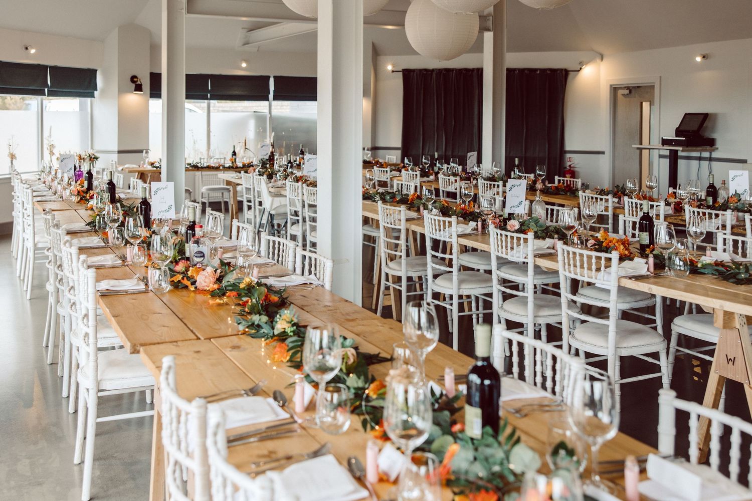 Modern wedding reception venue with long wooden tables, white chairs, and elegant floral centerpieces in a bright space.