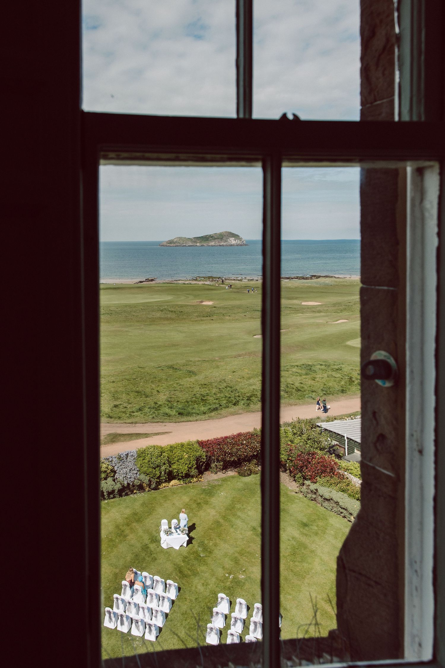 View through a window of white chairs set up for an outdoor wedding ceremony on a scenic coastal golf course.