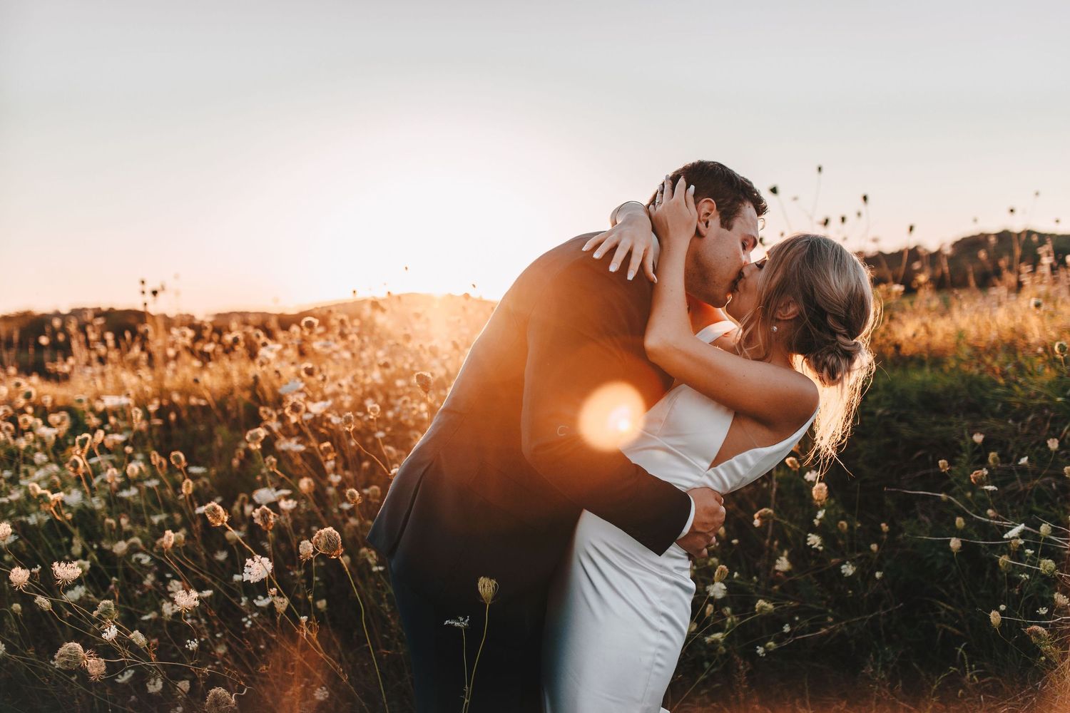 Bride and groom sharing romantic kiss in wildflower field during golden hour sunset.