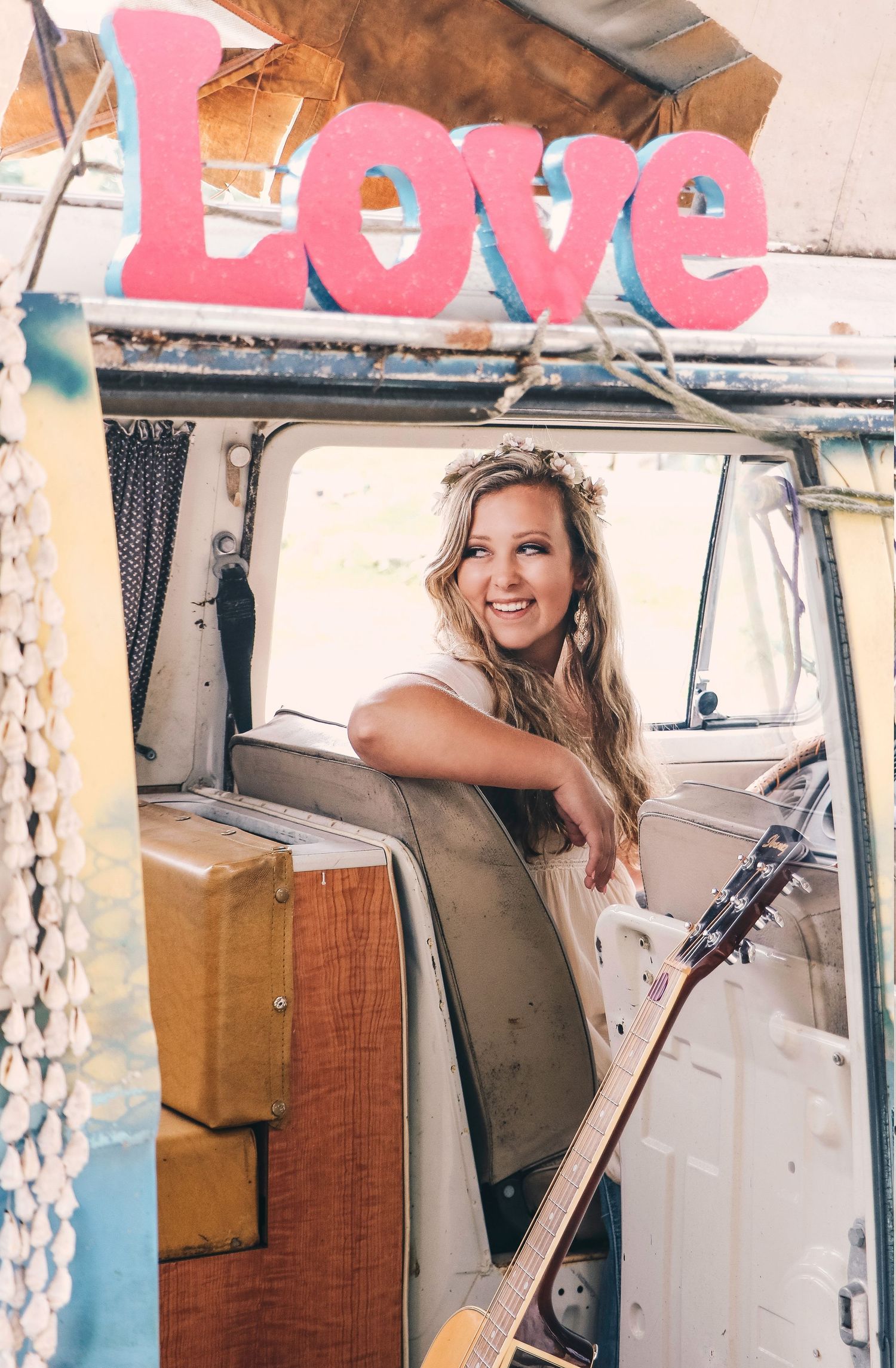 Smiling blonde woman with guitar leaning out of a vintage VW bus under a Love sign.