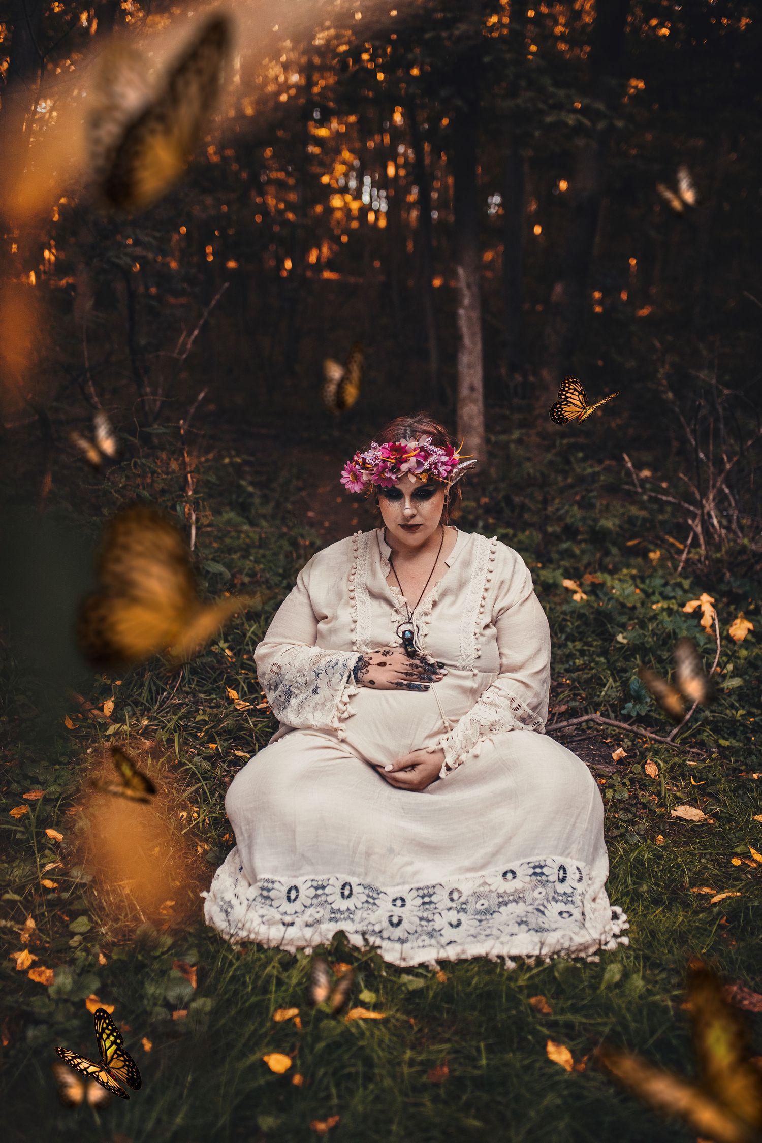 Woman in white dress with floral crown sitting among monarch butterflies in forest.