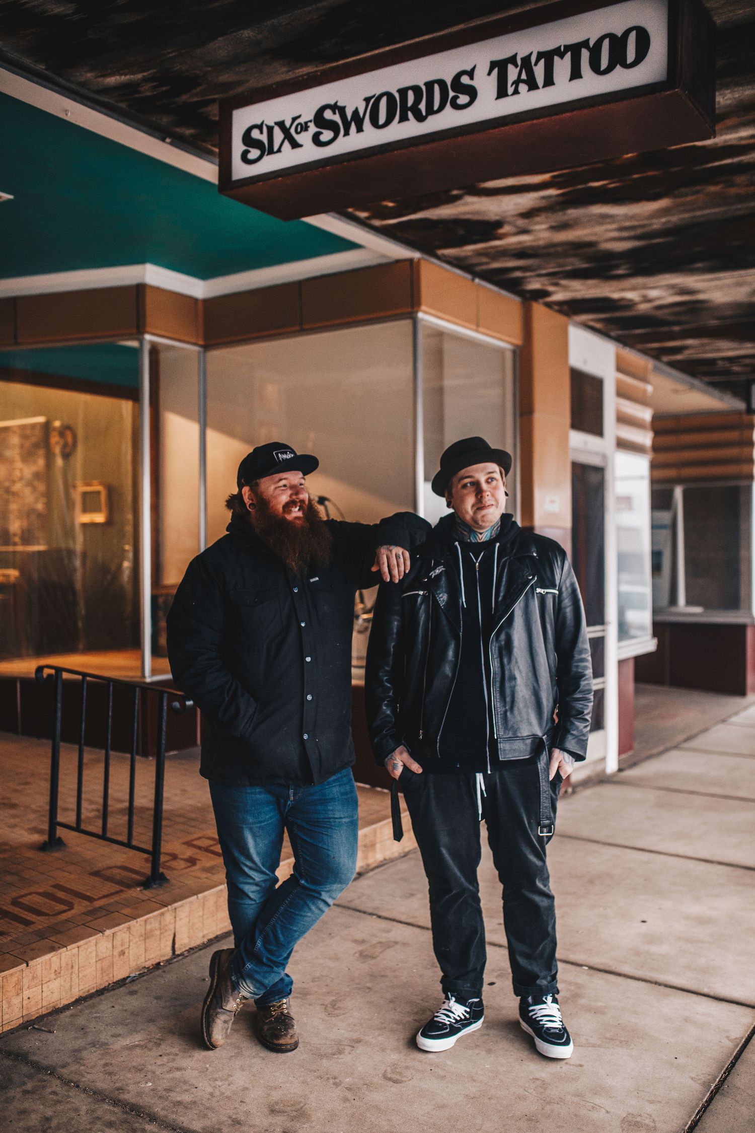 Two men posing outside Six Swords Tattoo shop on urban street.