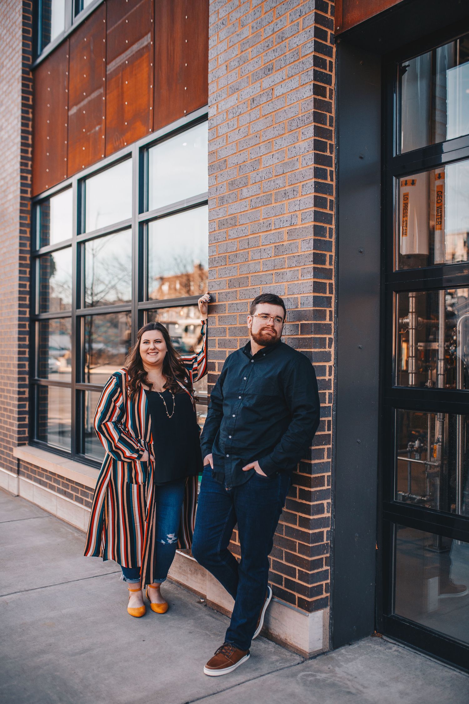 Couple posing against brick building exterior with modern black windows during golden hour engagement photos.