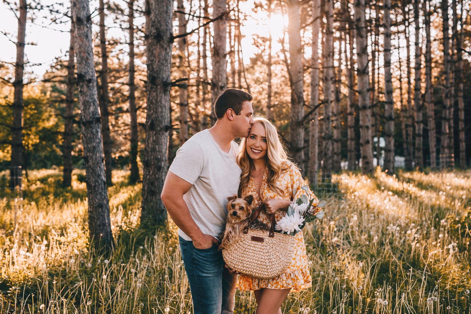 Couple embracing in sunlit forest at golden hour, woman holding bouquet of wildflowers.