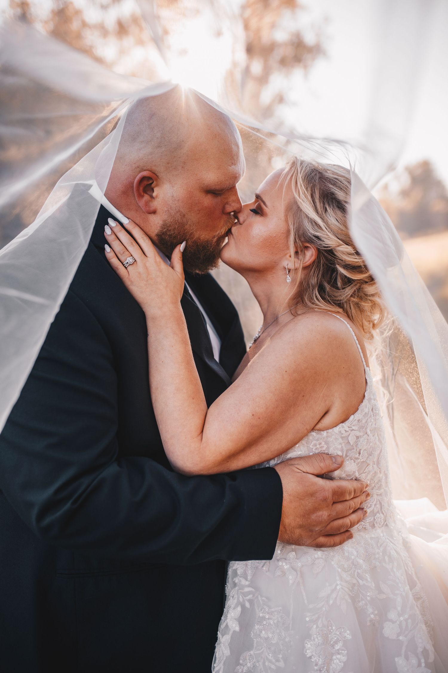 Bride and groom sharing romantic kiss under flowing veil in soft golden sunlight.
