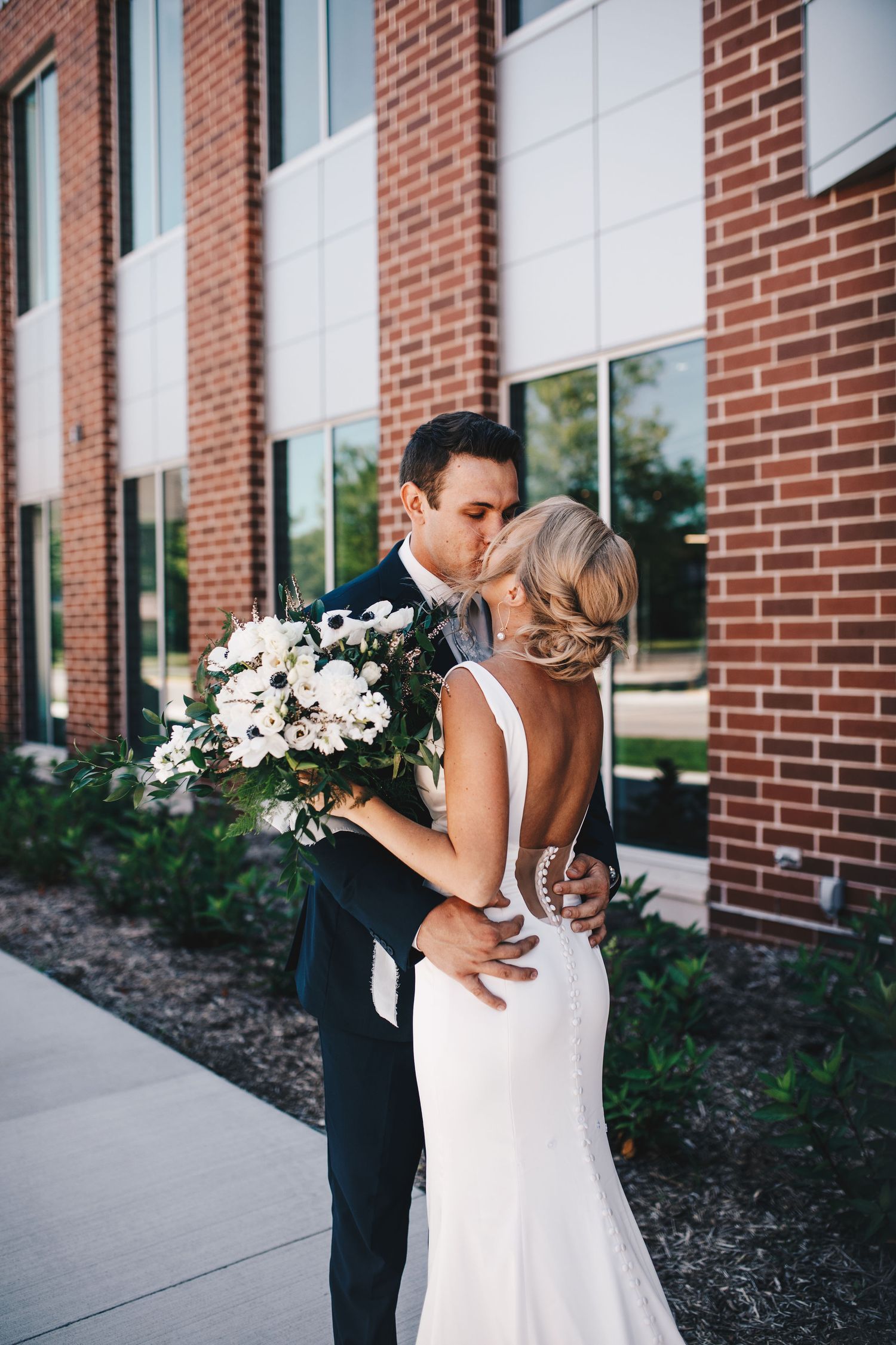 Groom in navy suit kisses bride in backless white gown while embracing near brick building exterior.