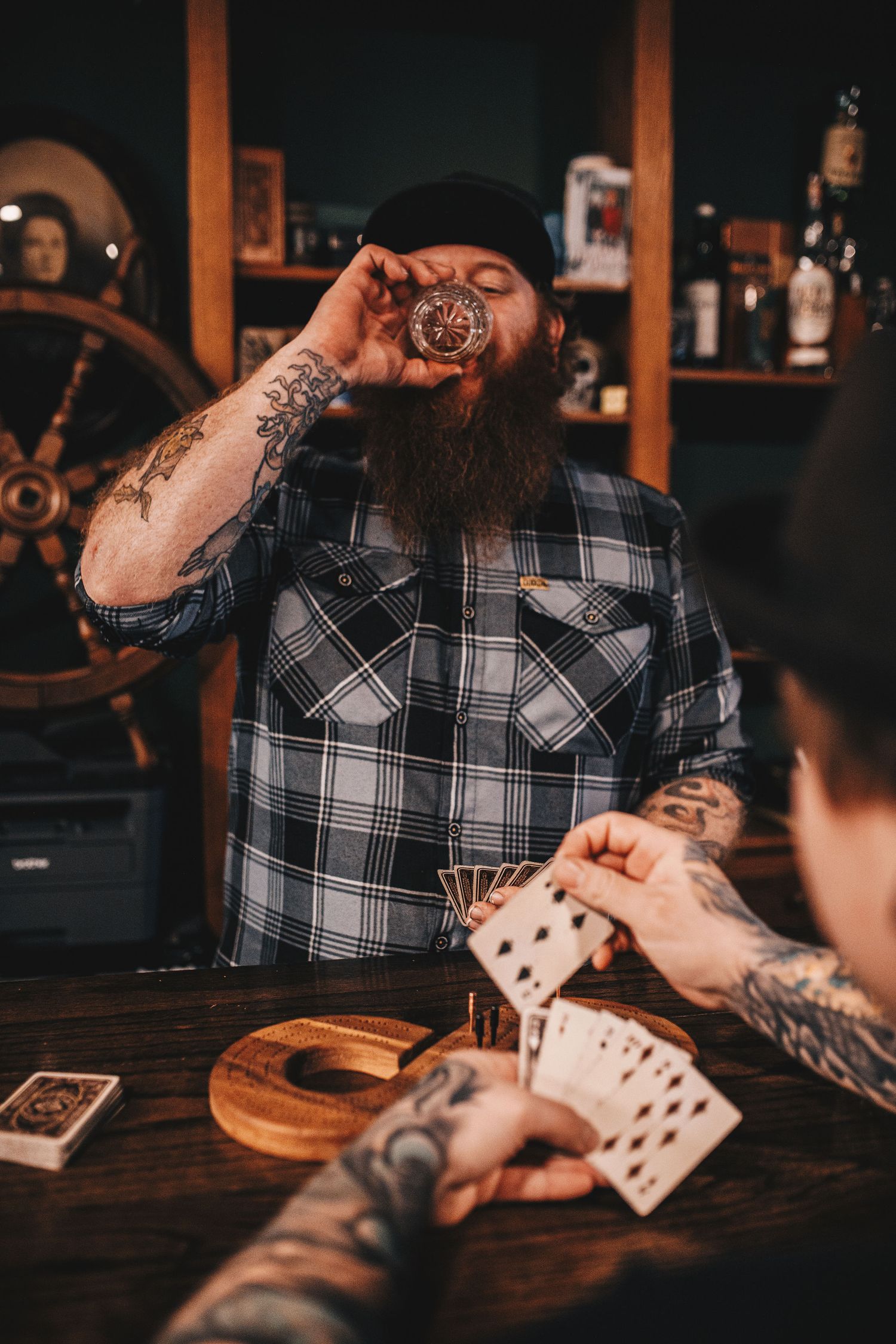 Bearded tattooed man drinking shot while playing cards at rustic bar.