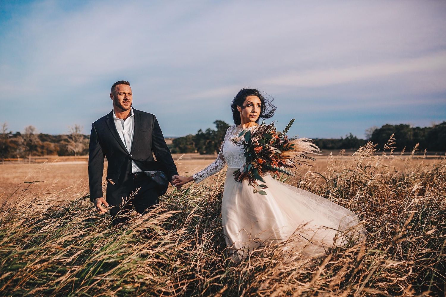 Bride in flowing white gown and groom walk through golden field under dramatic cloudy sky.