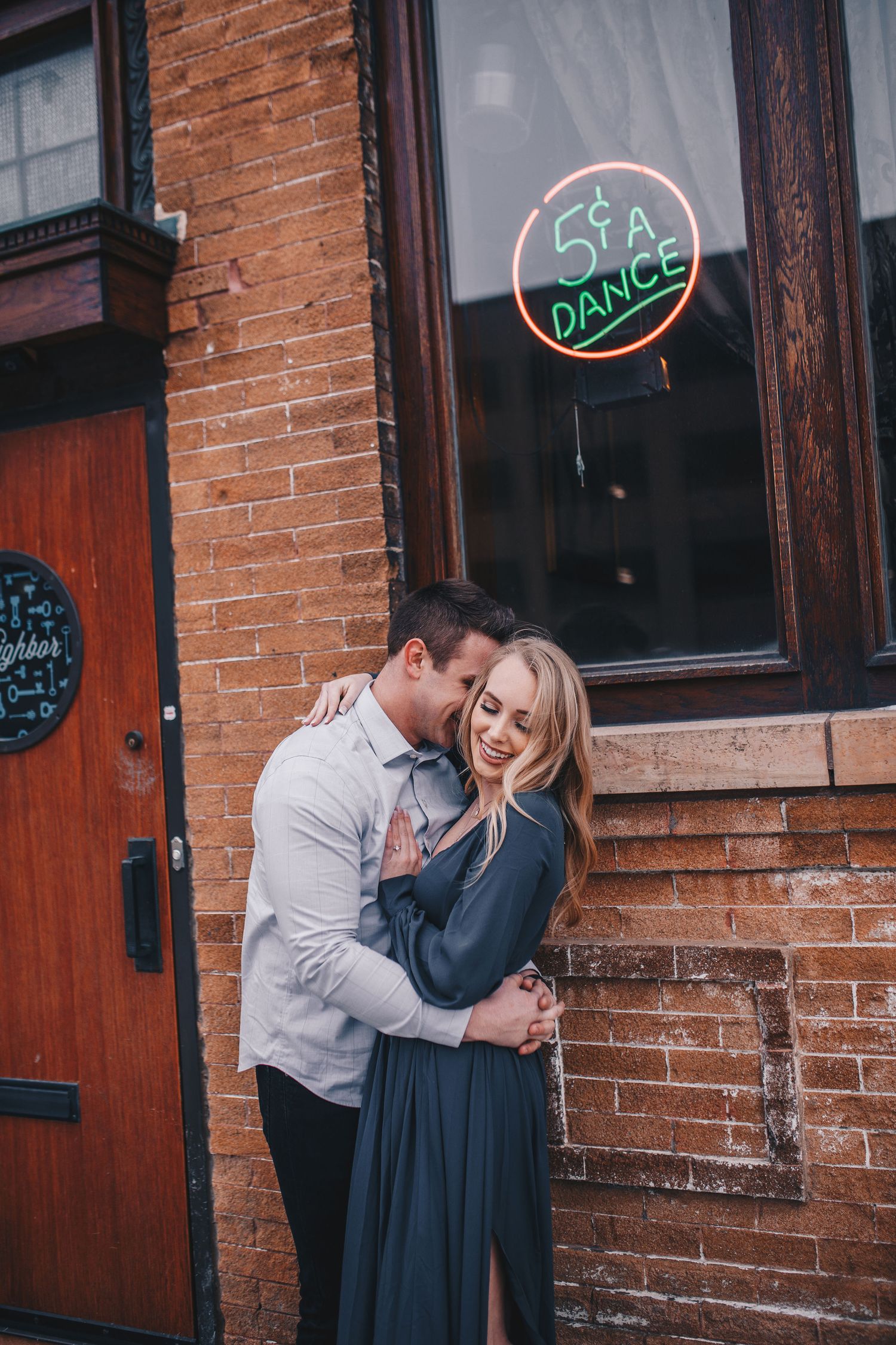 Romantic couple embracing and laughing outside vintage brick building with 5 cent dance neon sign.